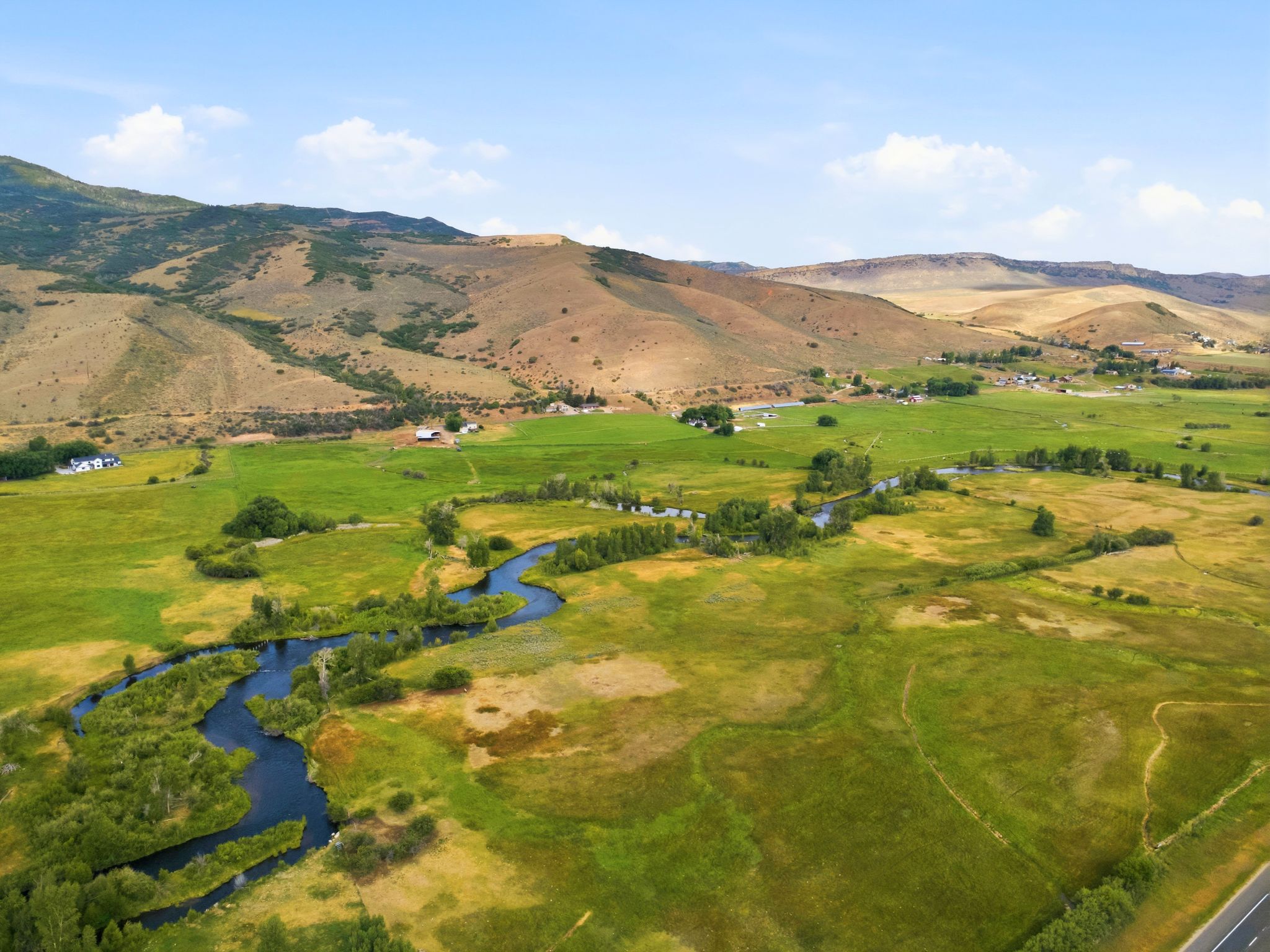 Expansive green and golden fields featuring a winding river, backed by rolling hills and distant structures