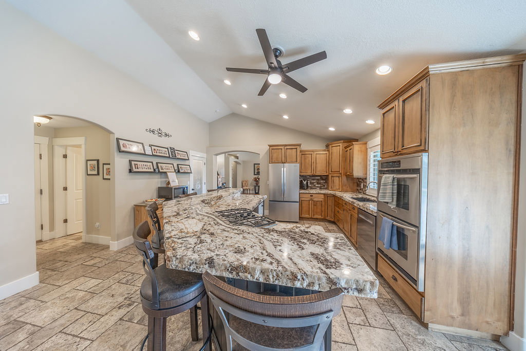 Spacious kitchen featuring an expansive island with a gas cooktop, natural wood cabinetry, stainless steel appliances including double ovens, vaulted ceilings with recessed lighting, and tile flooring