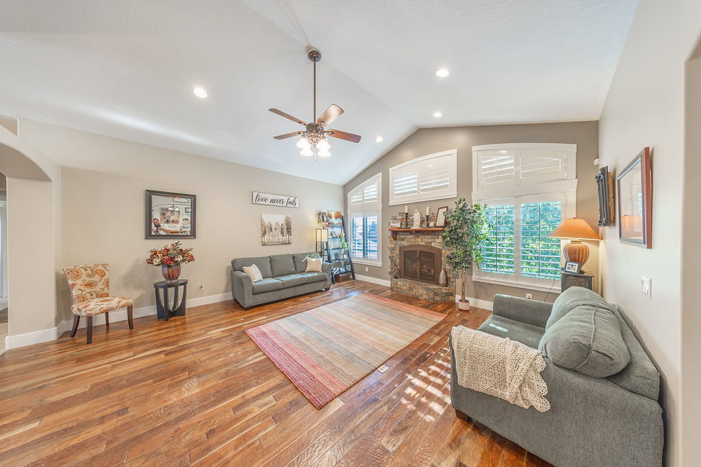 Spacious living area featuring vaulted ceilings with recessed lighting, wood-finish flooring, a stone-front fireplace, and multiple windows with plantation shutters
