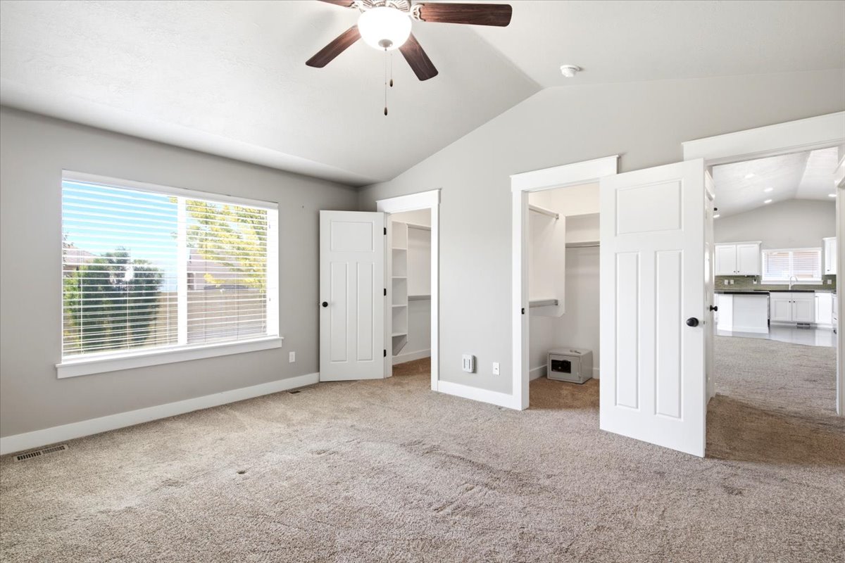 Carpeted room featuring a vaulted ceiling with a ceiling fan, a large window with blinds, and two open doorways revealing interior spaces