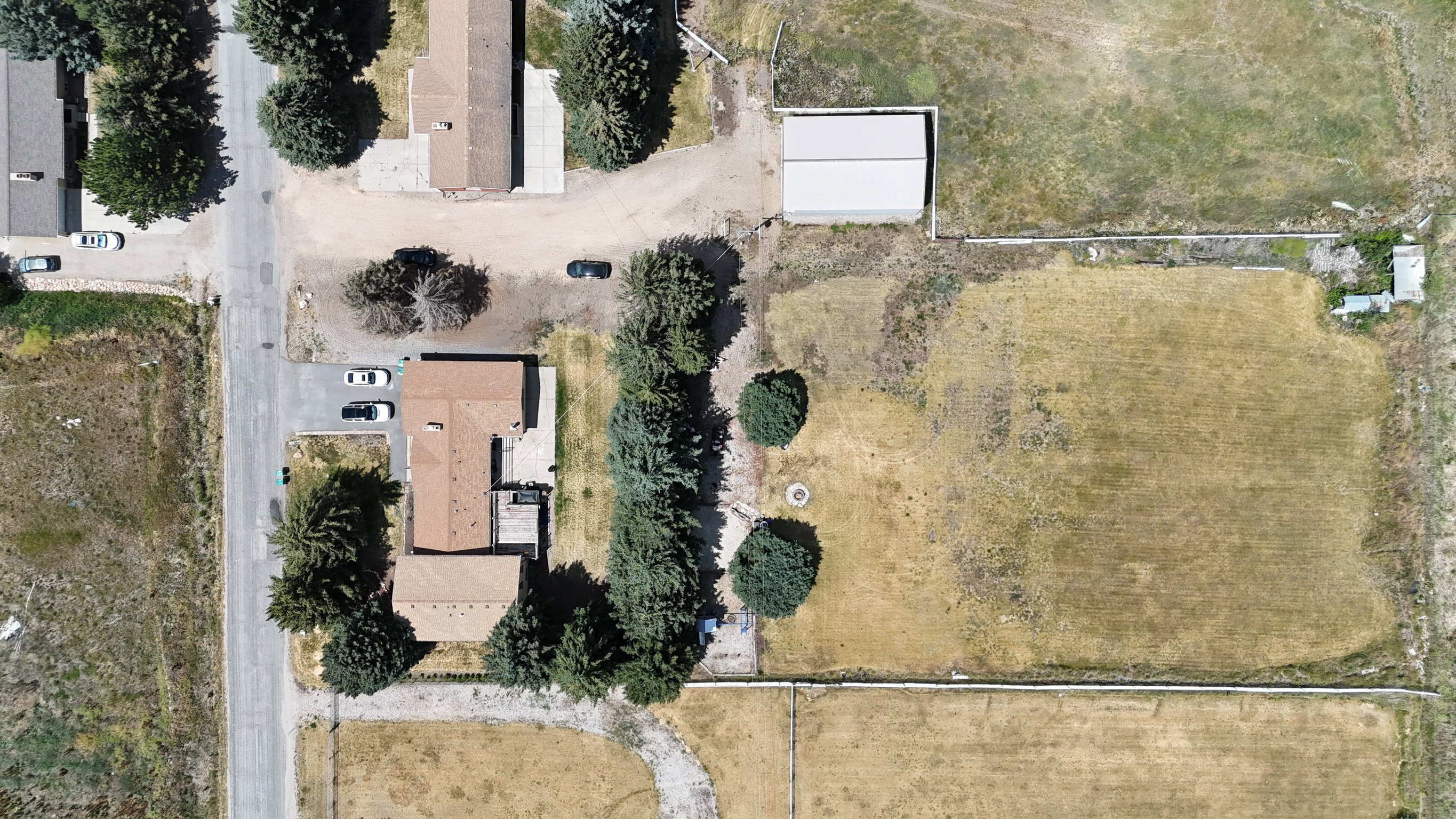 Expansive property featuring a brown-roofed residence, a substantial outbuilding with a light-colored roof, a paved road, and a large open field