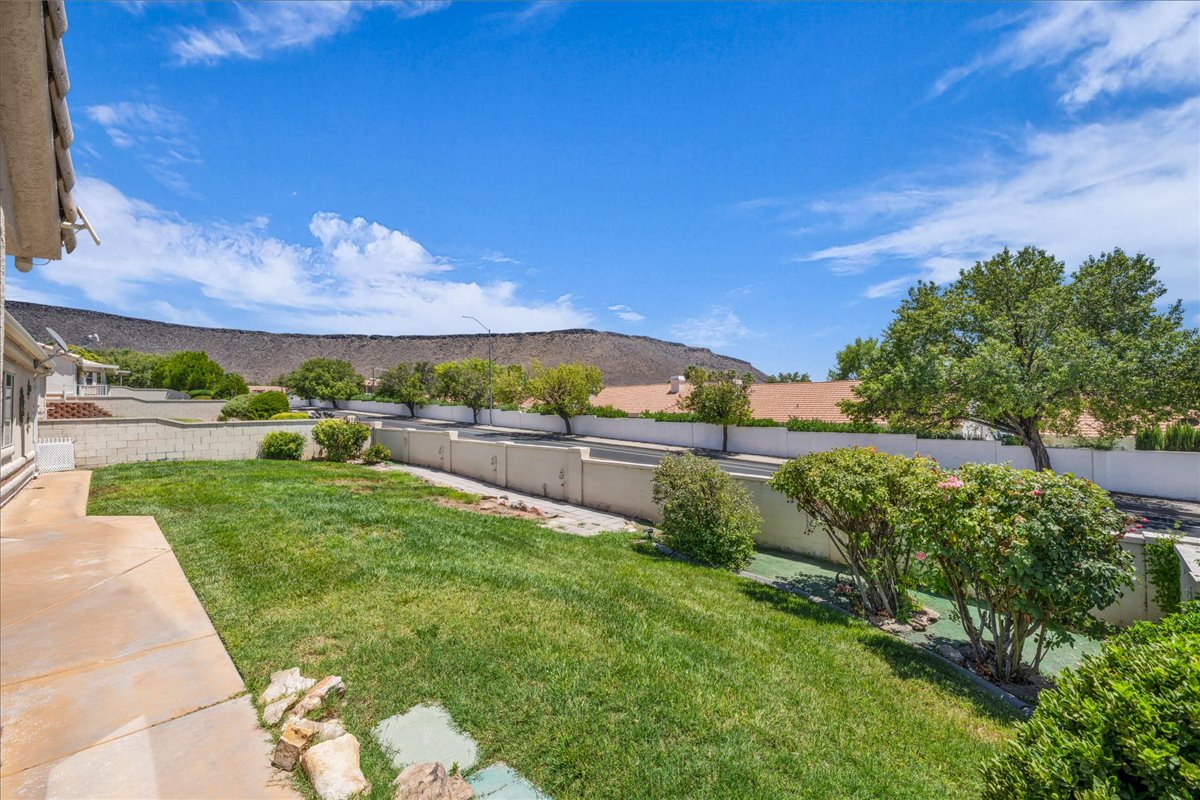 Spacious backyard featuring a large grass lawn, mature landscaping, a concrete patio, and a retaining wall, all set against a distant mountain backdrop