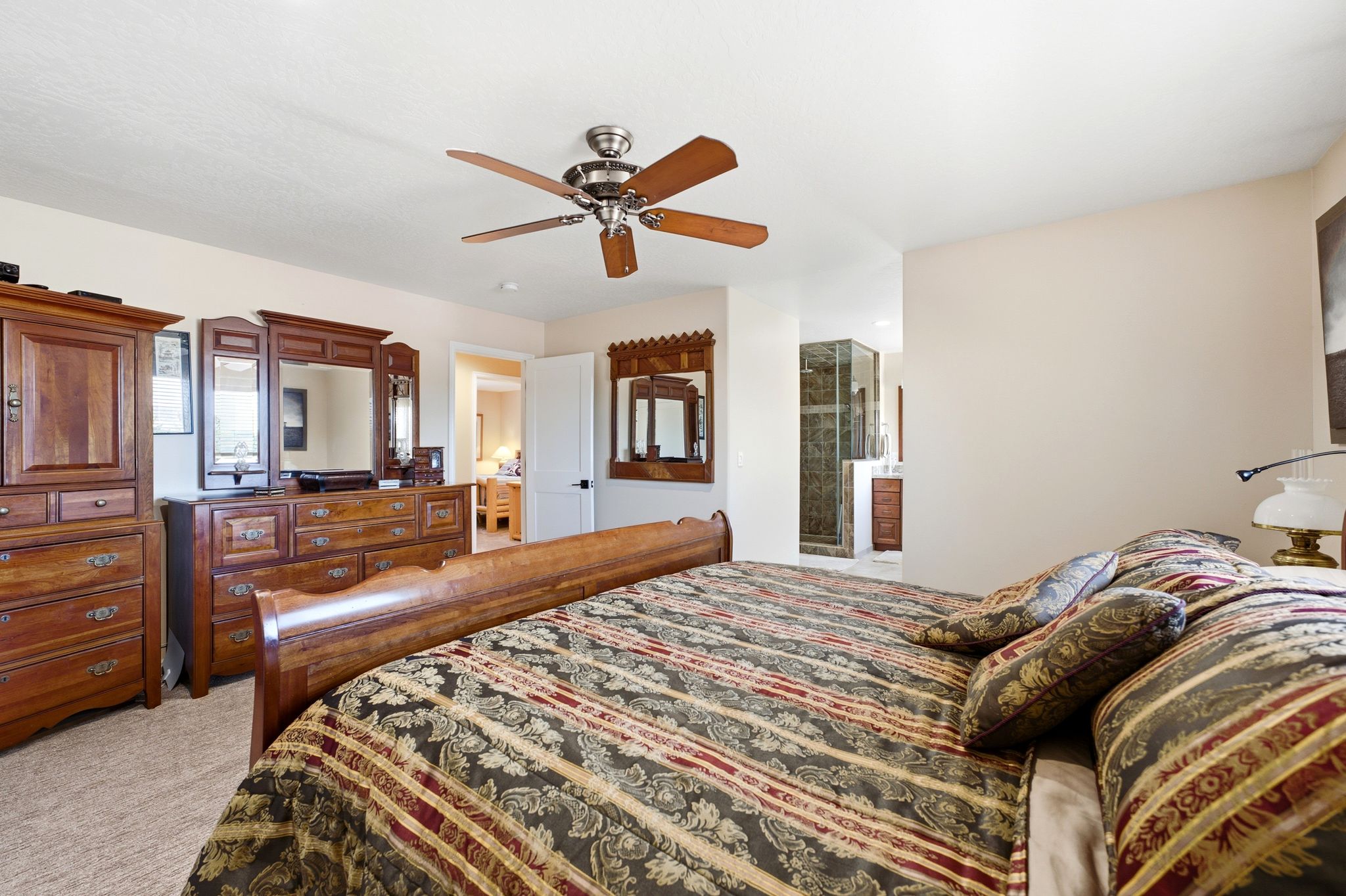 Carpeted room featuring a ceiling fan with wood-finish blades, a white door frame, and a partial view into an adjoining space with a glass-enclosed shower