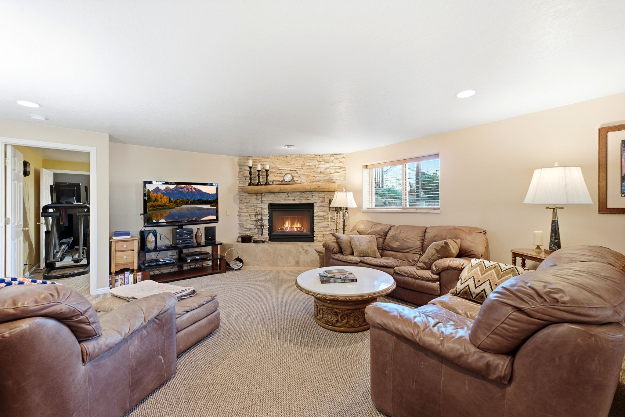 Spacious living area featuring a stacked stone fireplace with a natural wood mantle, recessed lighting, neutral carpeting, and light-toned wall finishes