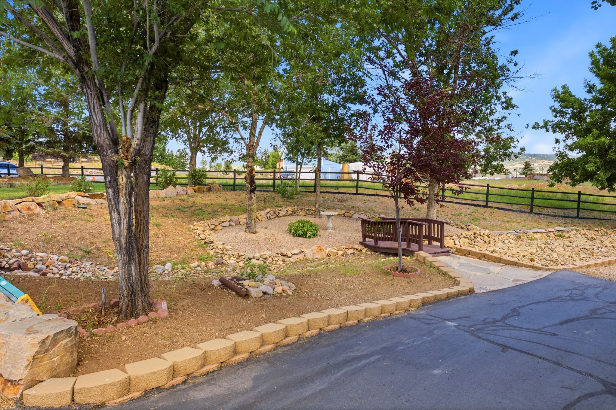 Landscaped yard featuring mature trees, a wooden footbridge, rock garden elements, and a paved driveway