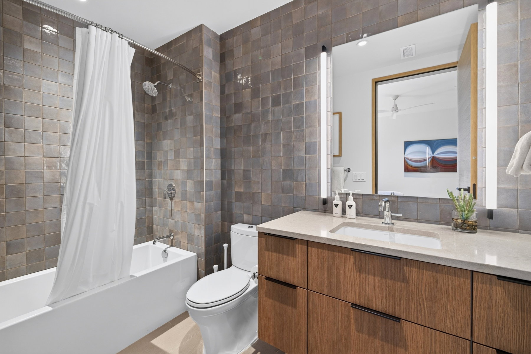 Contemporary bathroom featuring a dark tile shower surround, a white bathtub, and a modern vanity with wood-finish cabinetry, a light-toned countertop, and an integrated rectangular sink