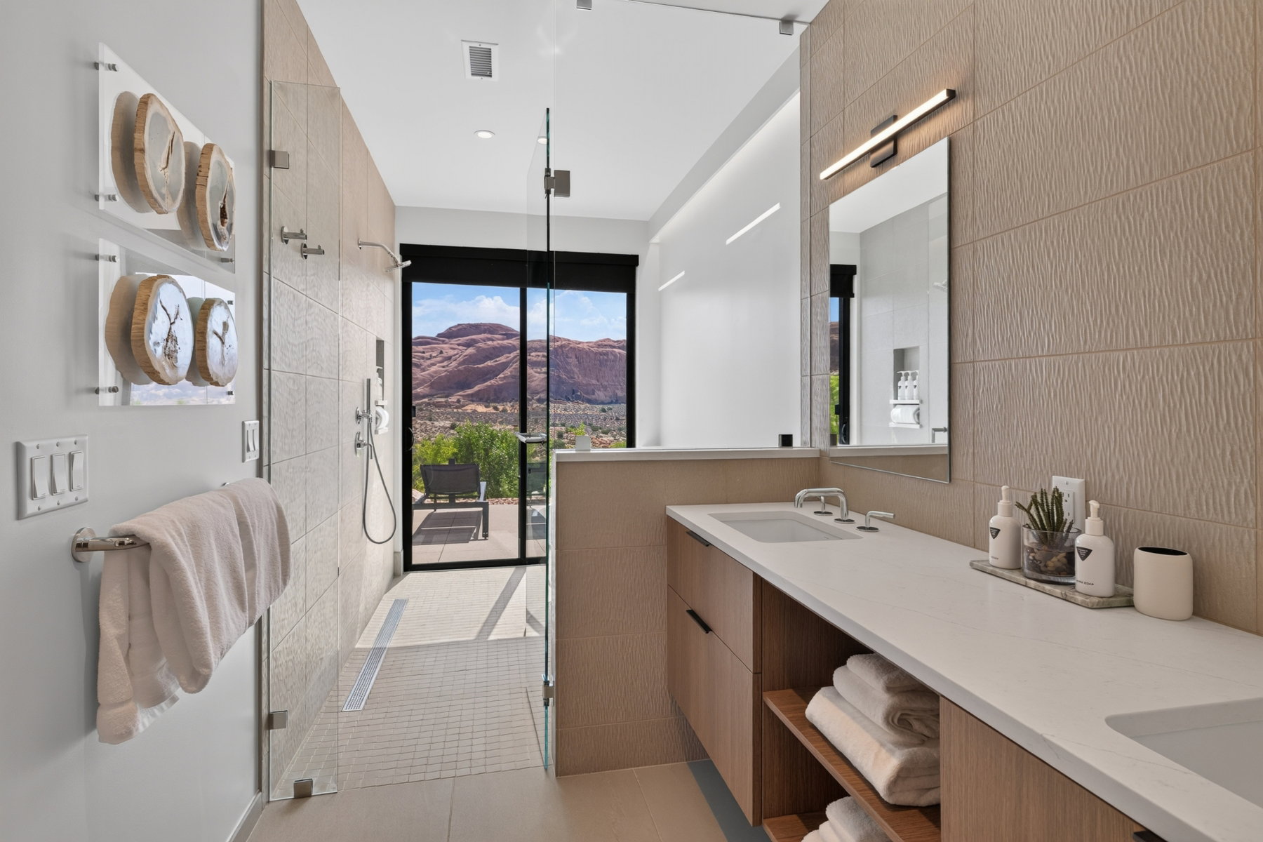 Contemporary bathroom featuring a dual vanity with light-tone wood-finish cabinetry, a large walk-in shower with glass enclosure, textured wall tiling, and a floor-to-ceiling window offering scenic natural rock formations