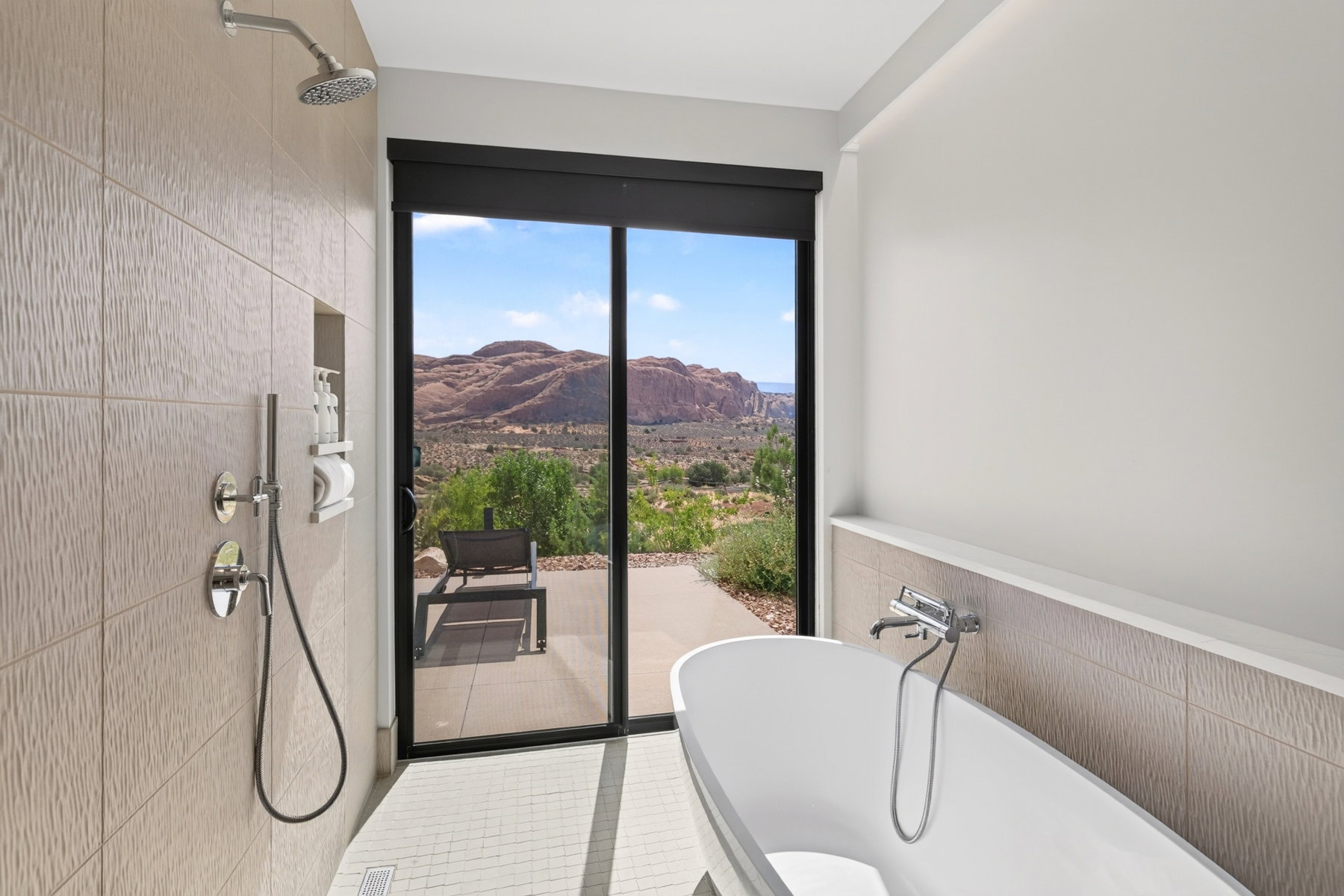 Freestanding soaking tub and shower area with textured tile, chrome fixtures, and glass sliding doors opening to a hardscaped patio