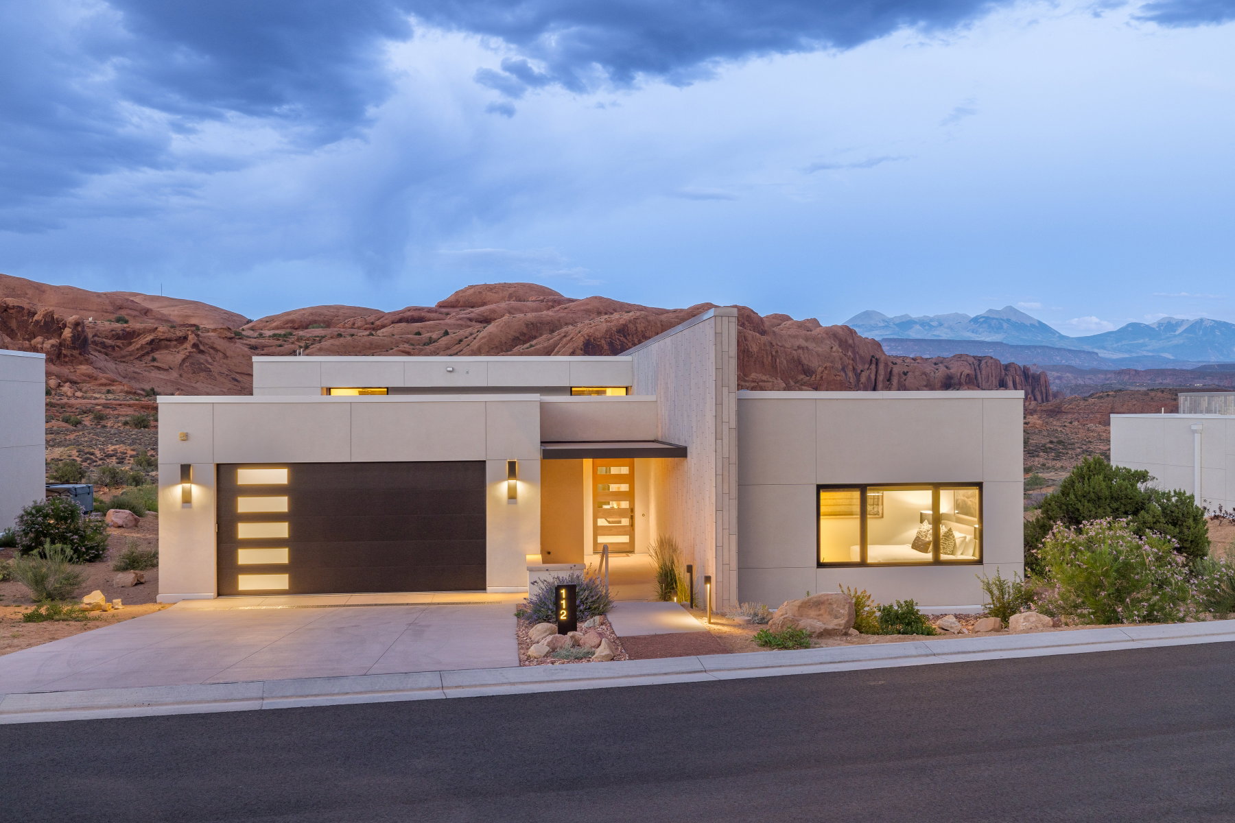 Modern architectural exterior with a flat roofline, integrated garage with frosted glass panels, illuminated entryway, and desert landscaping