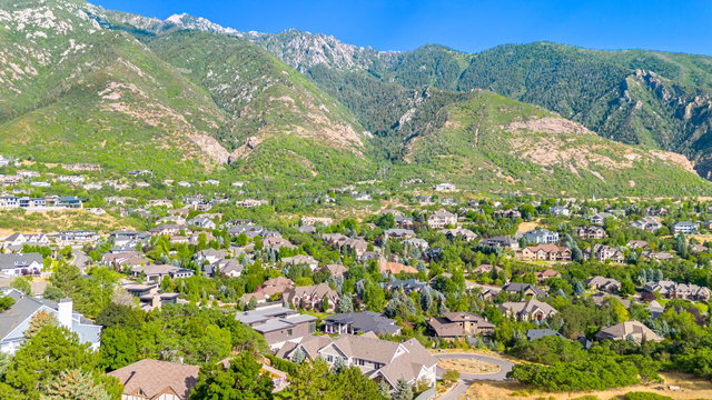 Residential neighborhood nestled at the base of verdant mountains, featuring various architectural styles, ample mature trees, and a clear blue sky