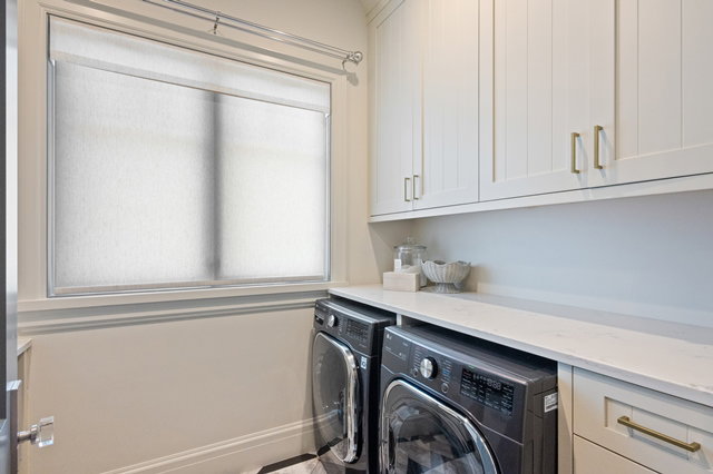 Laundry area featuring a window with roller shades, light-toned upper cabinetry with gold-finish hardware, a built-in countertop, and under-counter appliances