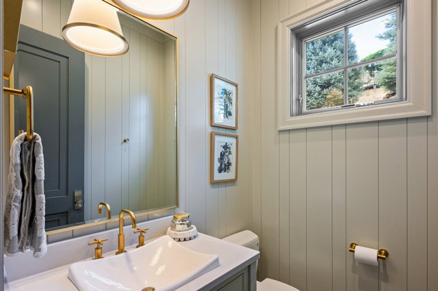 Board and batten wall paneling, vanity with a rectangular vessel sink, gold-finish faucet and hardware, two pendant light fixtures, and a window with exterior tree scenery