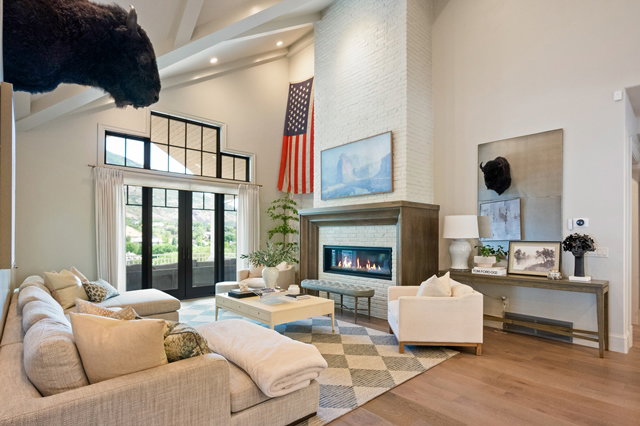 Spacious living area featuring a vaulted ceiling with exposed beams, light wood-finish flooring, and a floor-to-ceiling brick fireplace with a wood mantel
