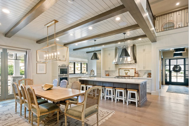 Open-concept interior featuring wood-finish flooring, a shiplap ceiling with exposed beams, a kitchen island with seating, a built-in oven, and a balcony with an iron railing