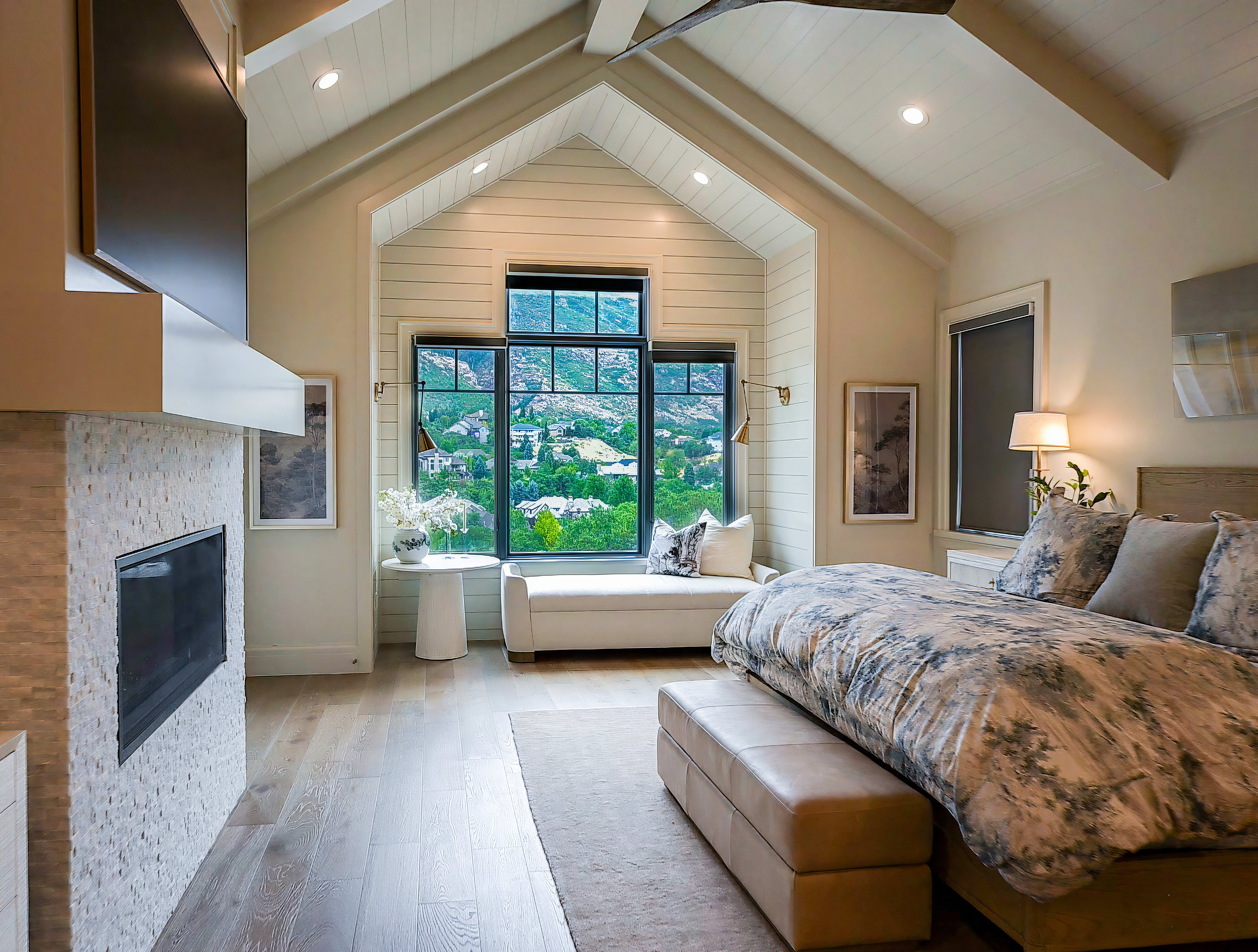 Vaulted ceiling bedroom featuring a floor-to-ceiling window, an integrated window seat, wood-finish flooring, a stone-clad fireplace with a mounted television, and recessed lighting