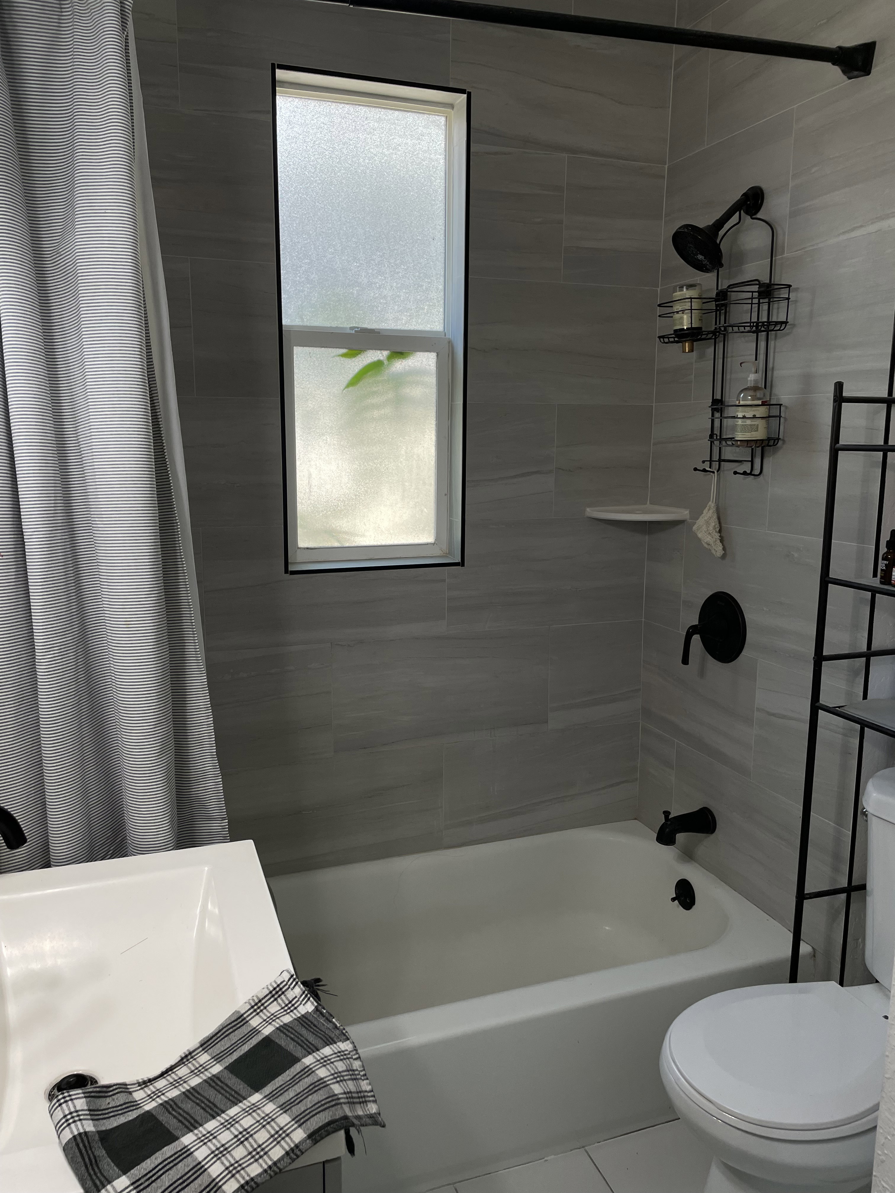 Bathroom featuring light gray tiled shower surround, a white bathtub, black fixtures, and a rectangular frosted window
