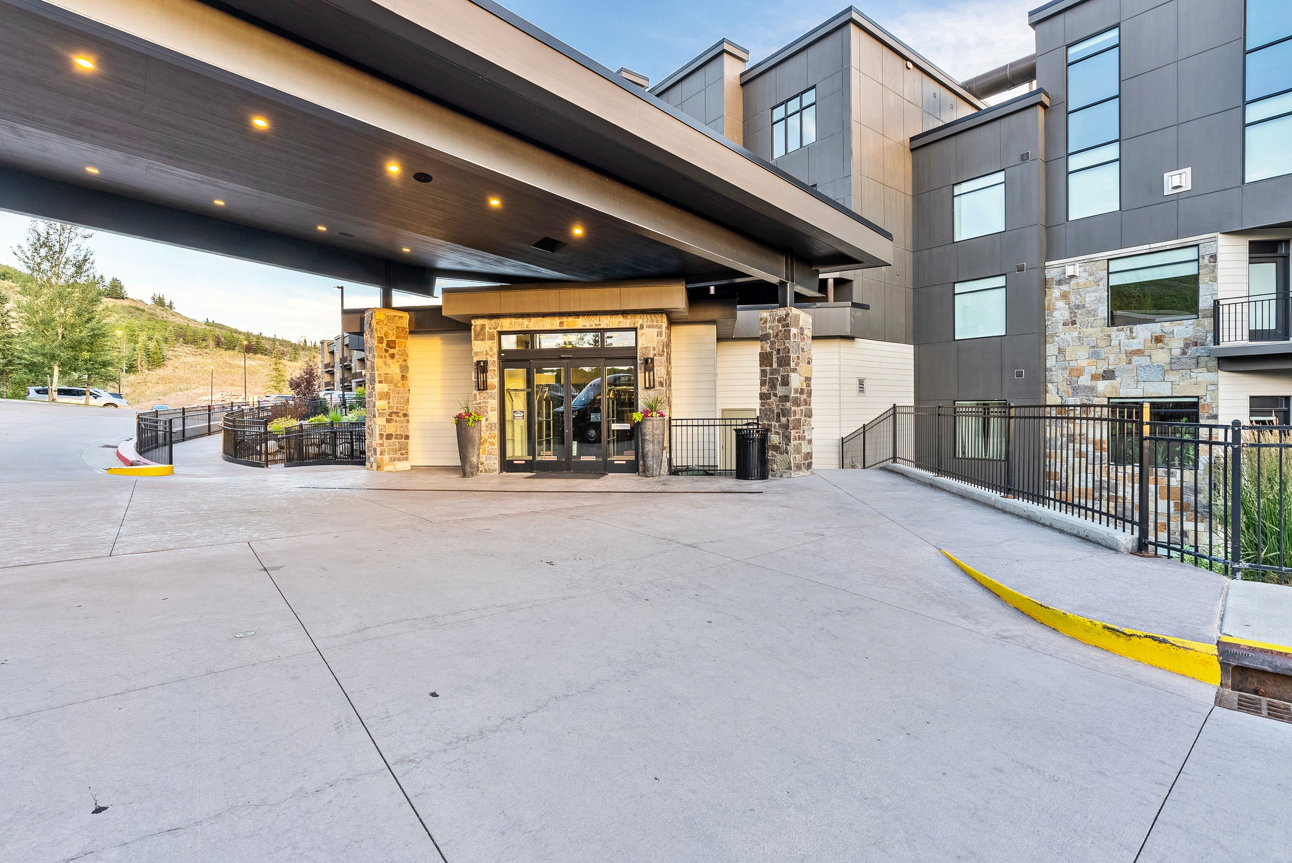 Modern building entrance featuring a covered drop-off area with recessed lighting, stone-clad pillars, and a glass-paneled double door entry