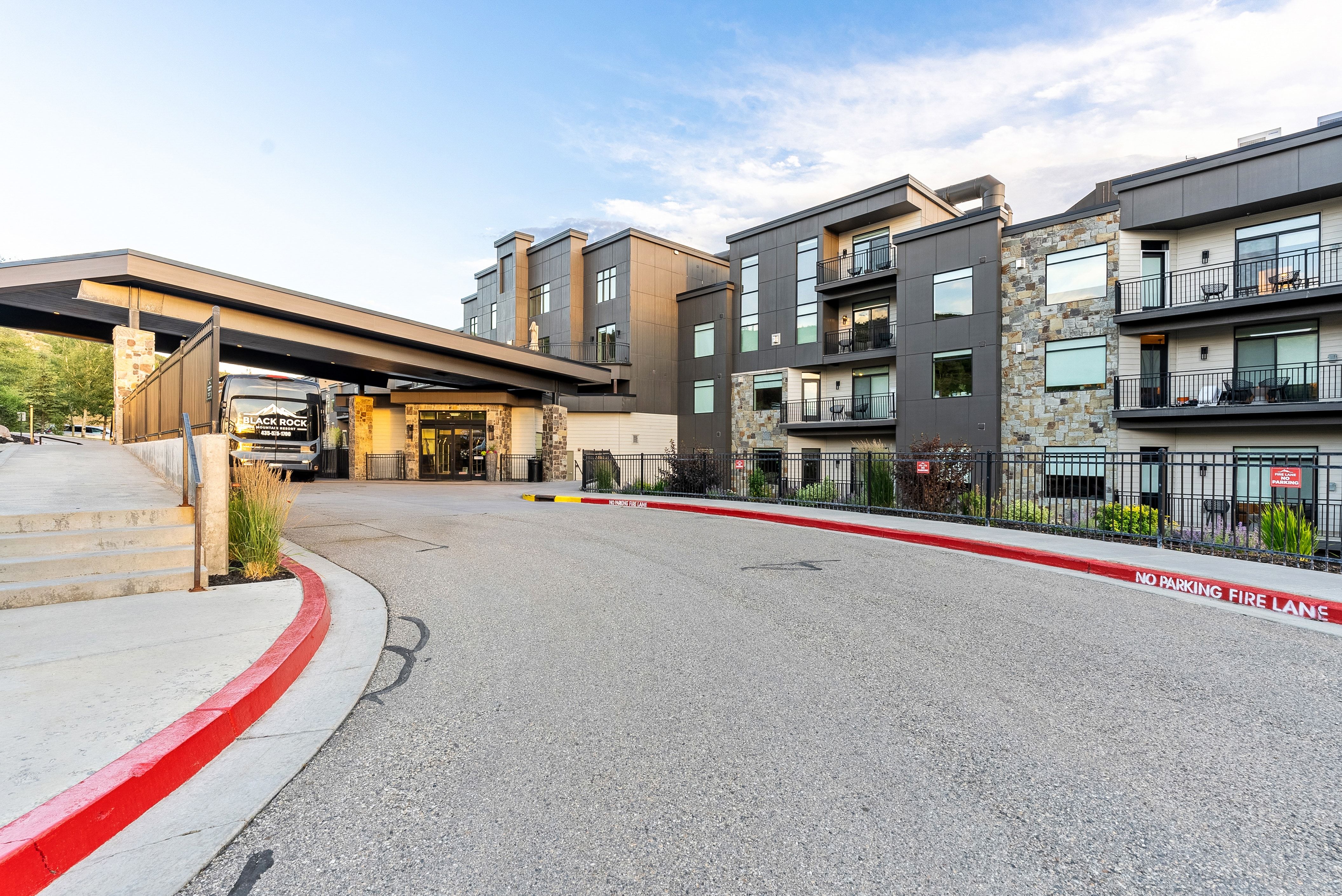 Modern multi-story building featuring stone and paneling facade, multiple private balconies with railings, covered entryway with stone columns, and an asphalt drive with concrete steps