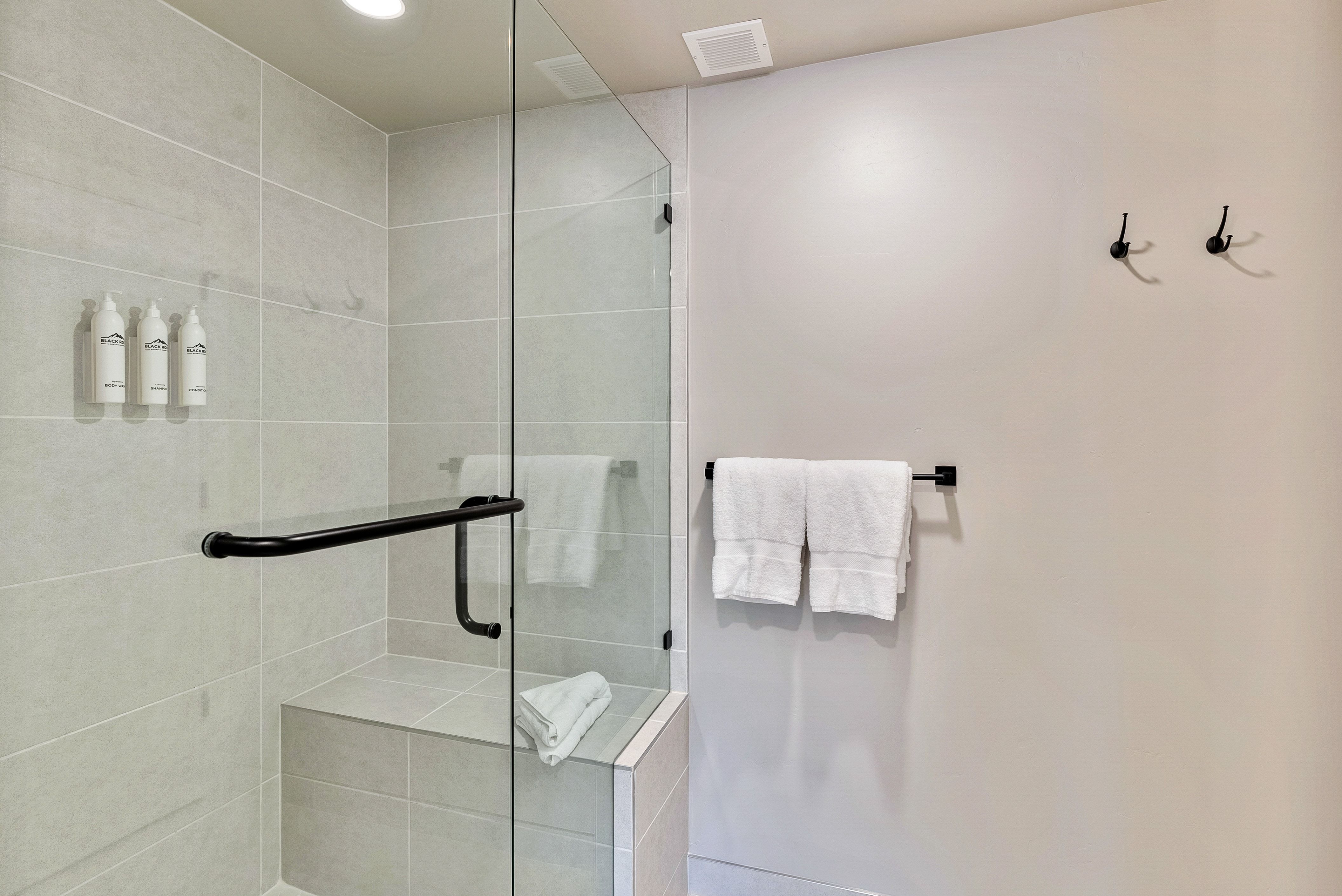 Glass-enclosed shower featuring light grey rectangular wall tiles, a built-in shower bench, a black shower handle, and a black grab bar