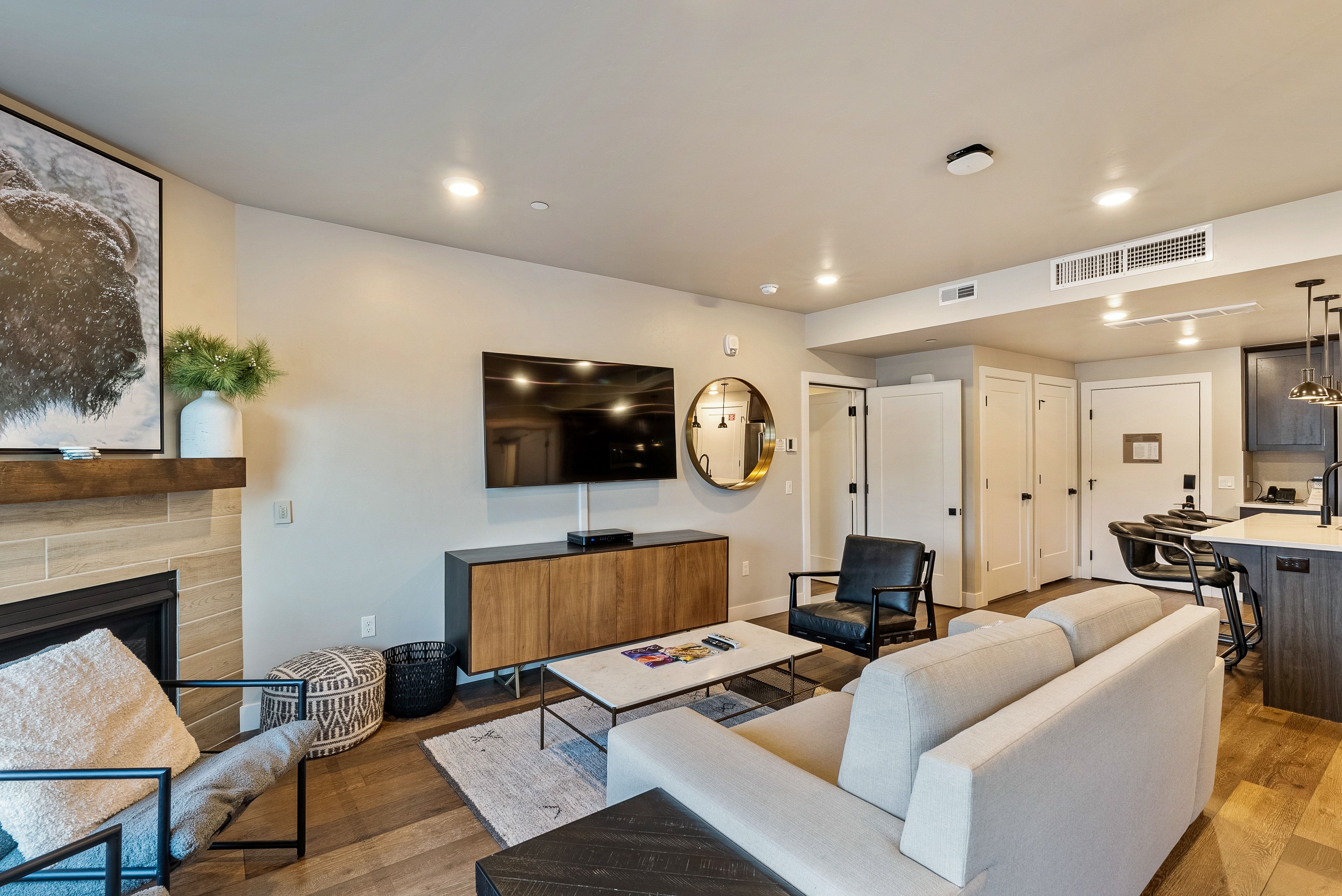 Open-concept living area featuring wood-finish flooring, a contemporary fireplace with a wood mantle, recessed lighting, and a kitchen island with pendant lighting