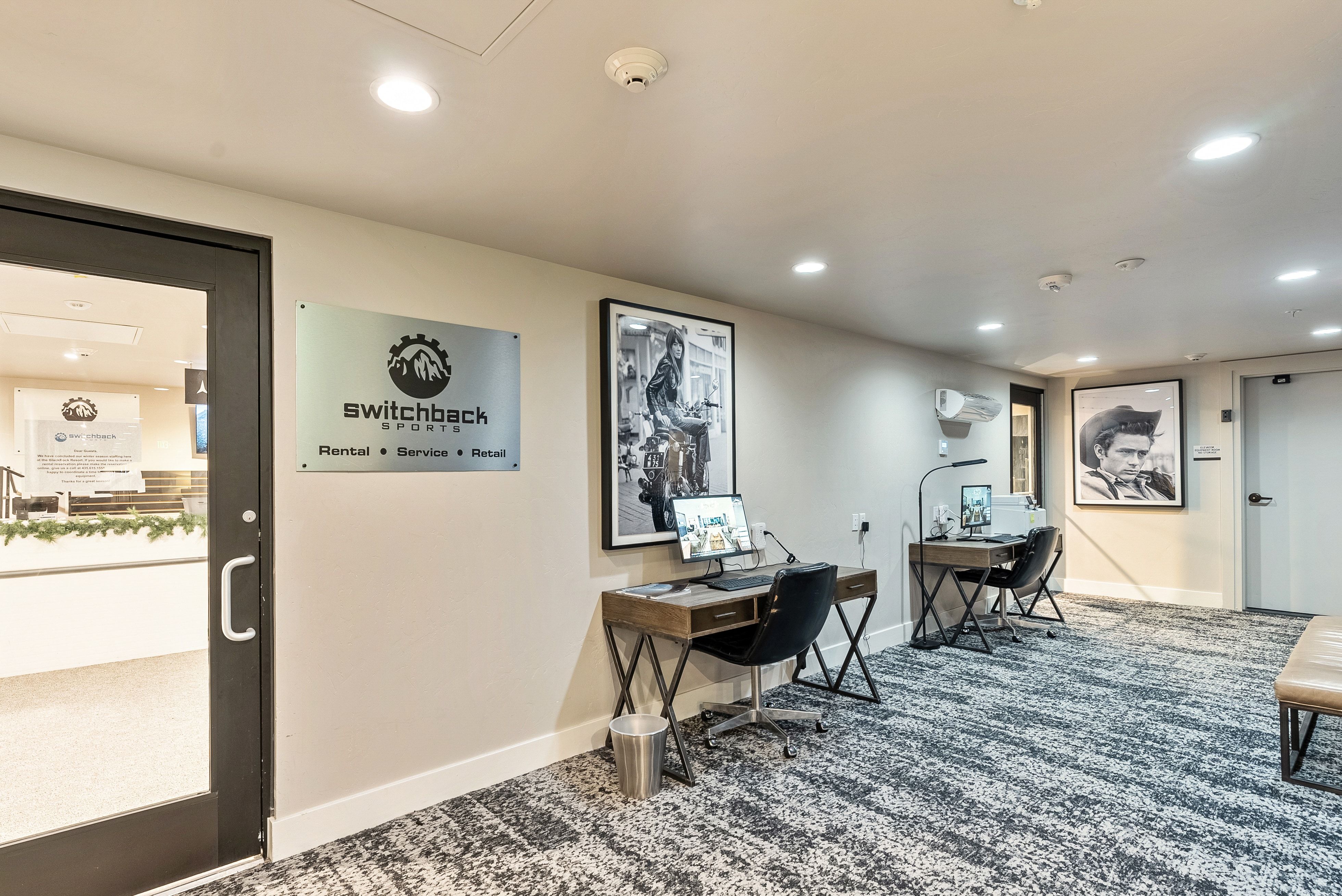 Lobby area featuring two workstation desks with monitors, a modern abstract patterned carpet, recessed ceiling lighting, and a frosted glass entry door