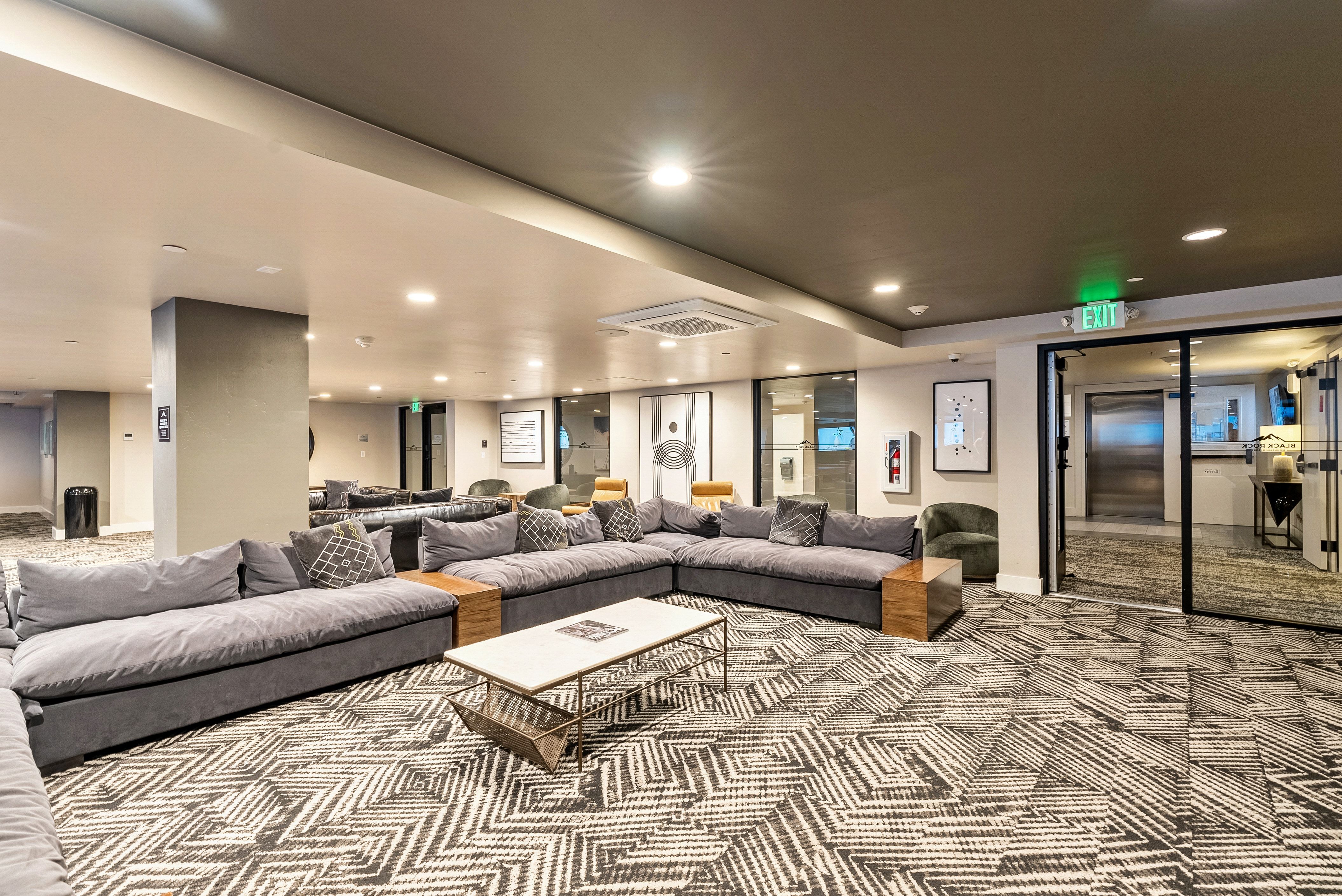 Spacious lounge area featuring patterned carpeting, recessed lighting, expansive sectional seating with wood-finish armrests, and glass panel doors