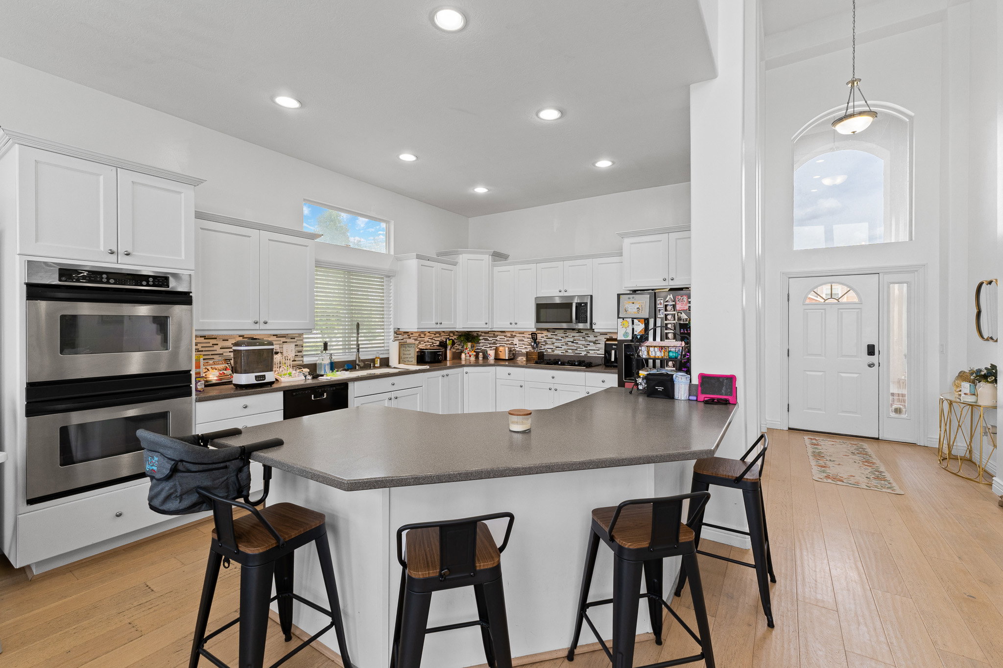 Open-concept kitchen featuring an eat-at island, stainless steel double ovens, wood-finish flooring, white cabinetry, and a full backsplash