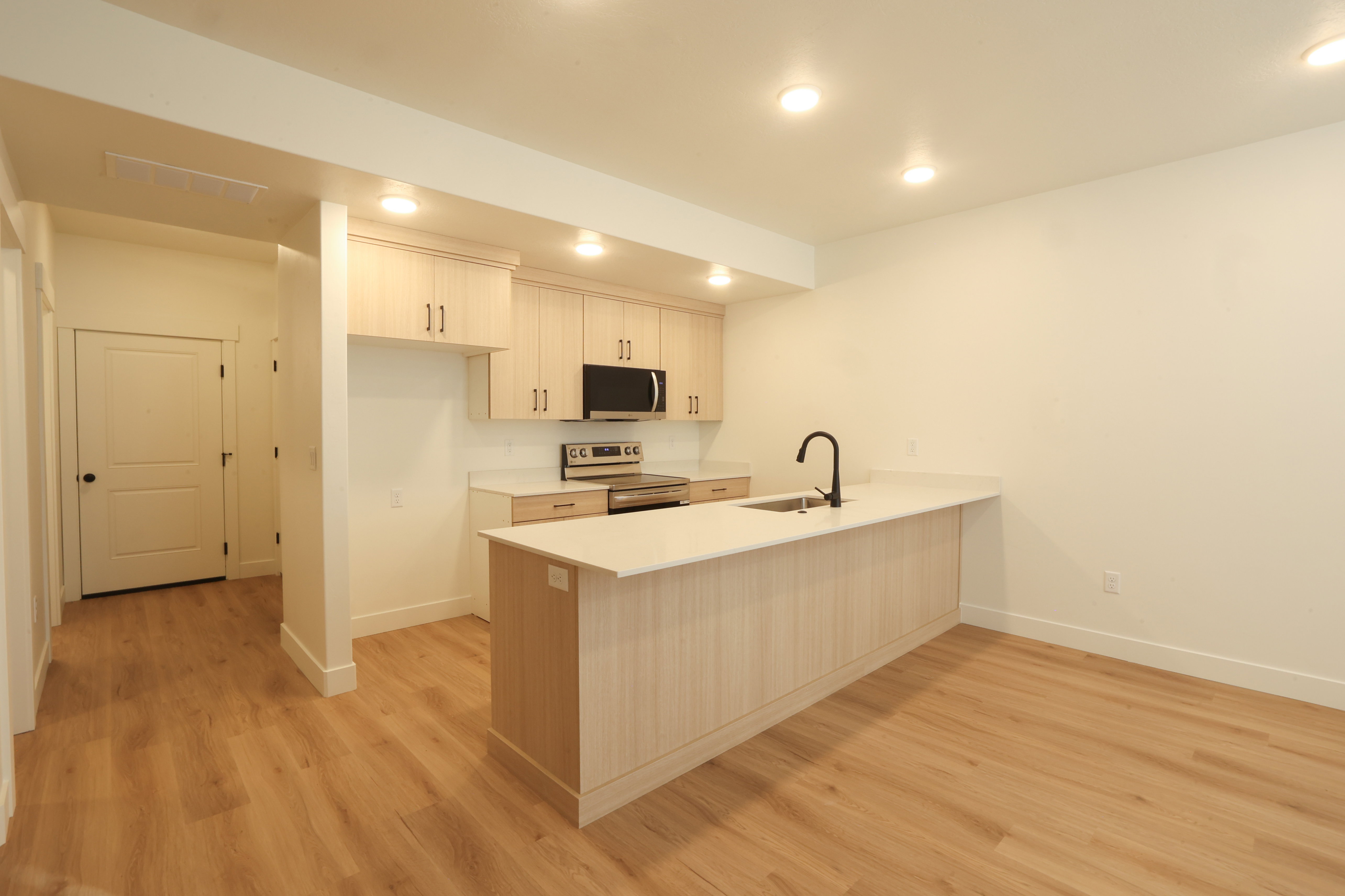 Open-concept kitchen featuring light wood-finish cabinetry, a large center island with a sink and matte black faucet, recessed lighting, and wood-finish flooring