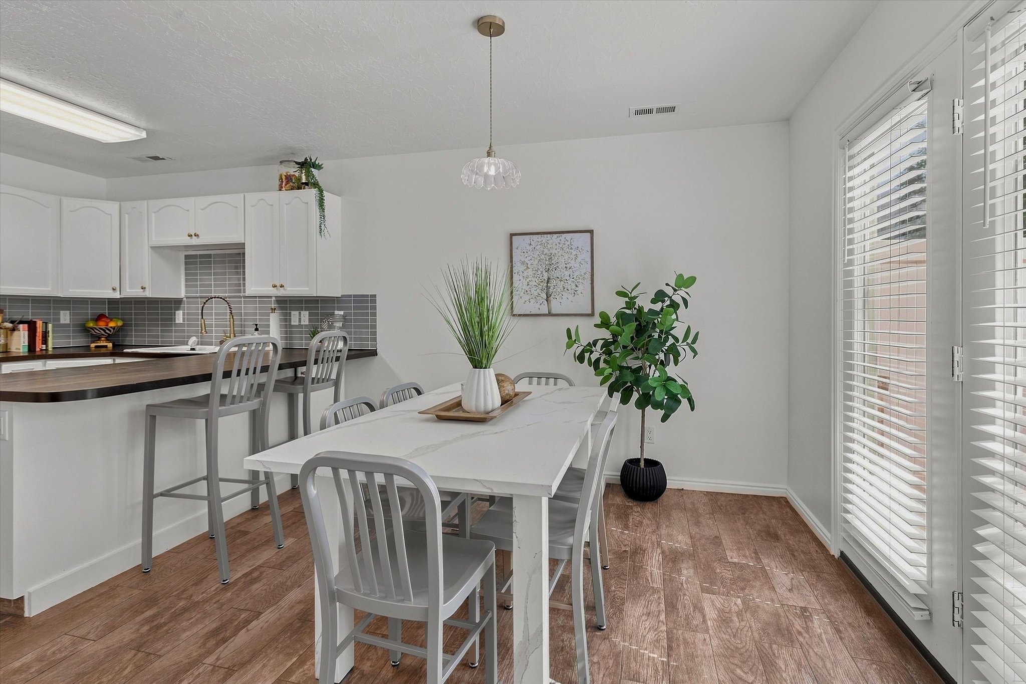 Open-concept dining area featuring wood-finish flooring, a white dining table with coordinating chairs, and a glass-paneled exterior door with blinds