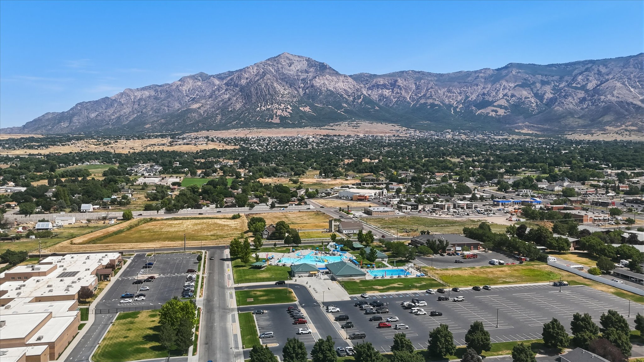 Expansive aerial capturing a mountain backdrop, a community aquatic center with multiple pools, and mature trees throughout the surrounding area