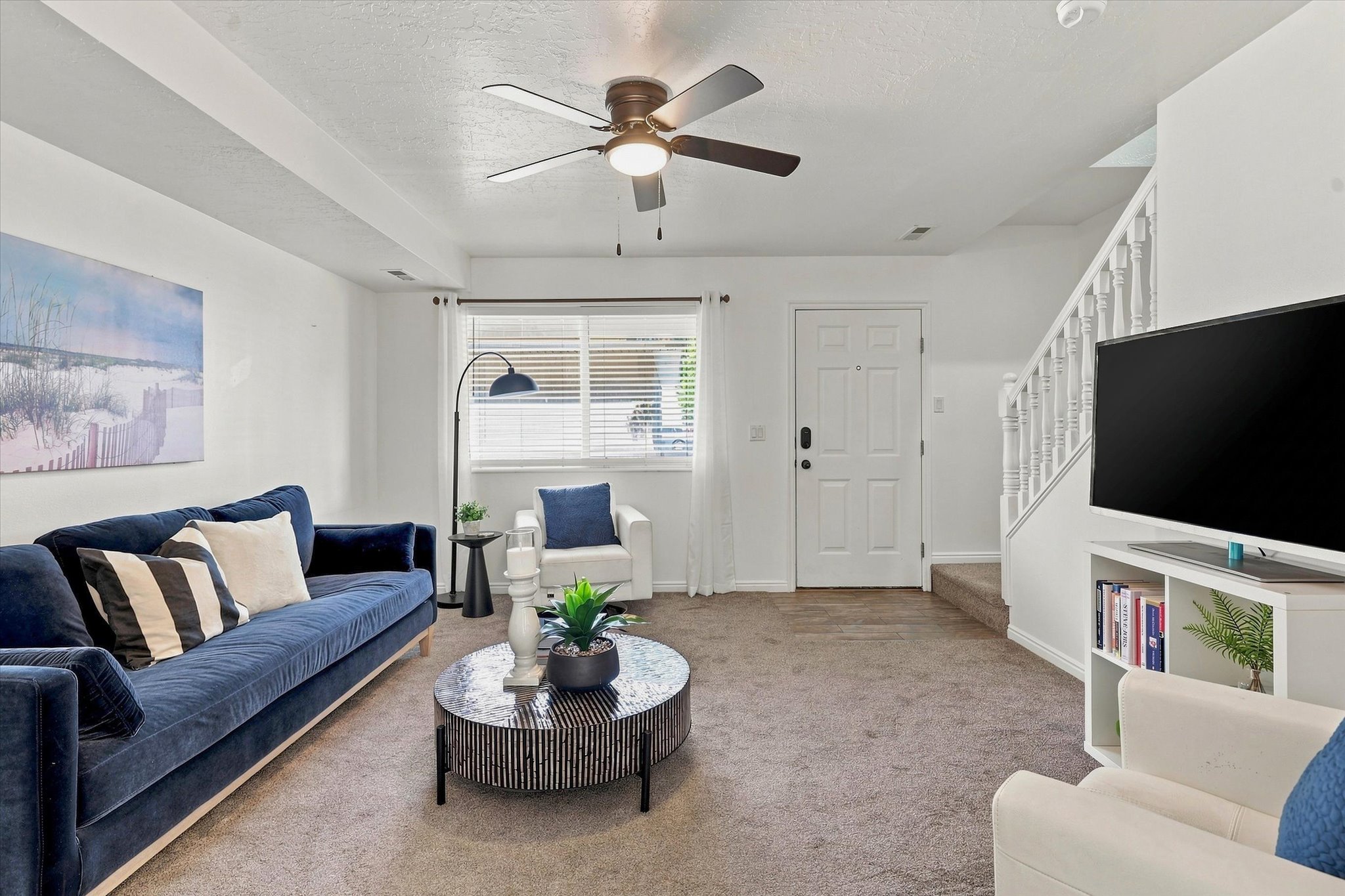 Living area featuring a textured carpet, a ceiling fan with integrated lighting, a white paneled entrance door, and a staircase with white balusters