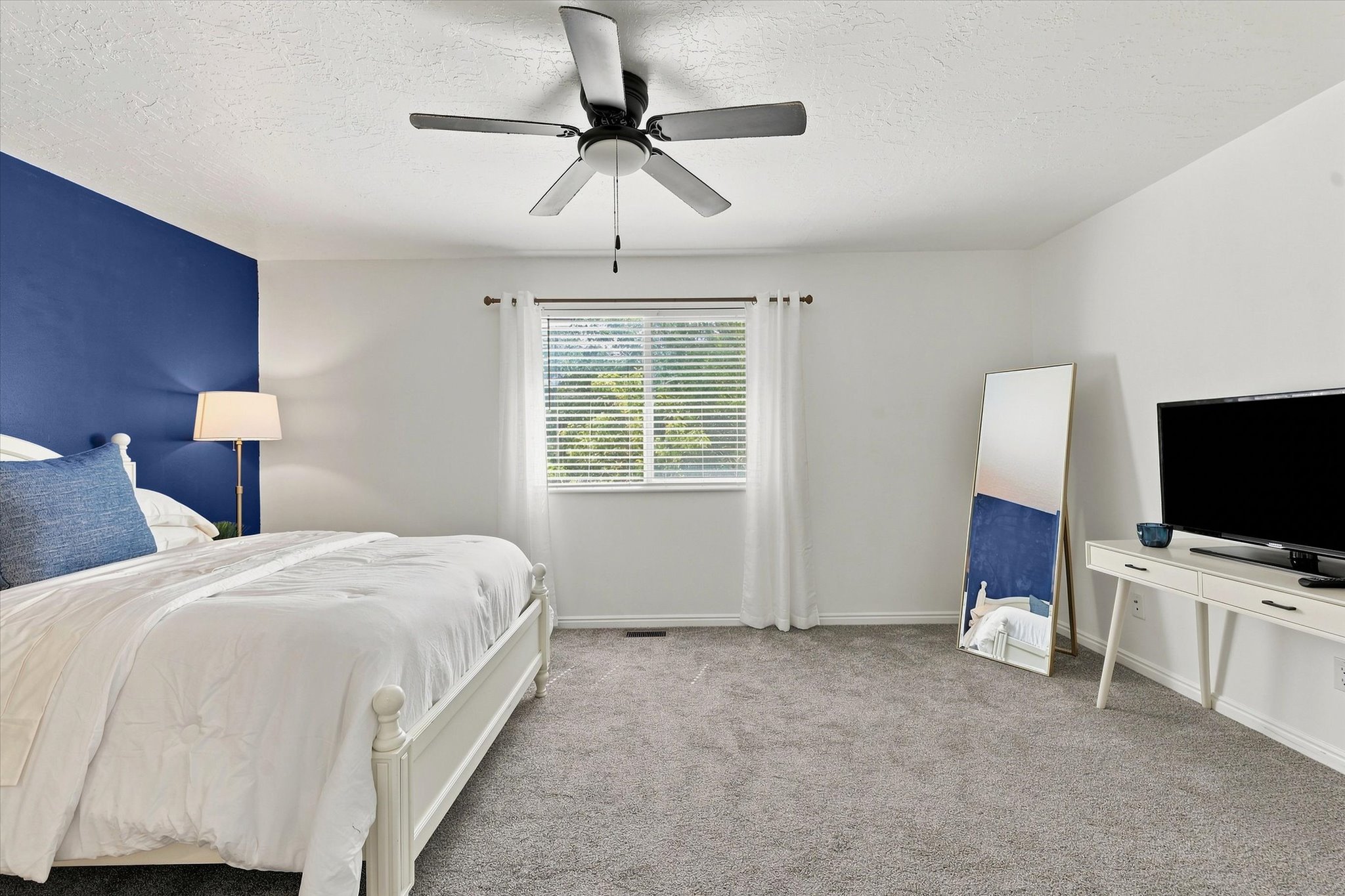 Spacious room featuring plush gray carpeting, a window with horizontal blinds and sheer white curtains, and a ceiling-mounted fan with integrated lighting