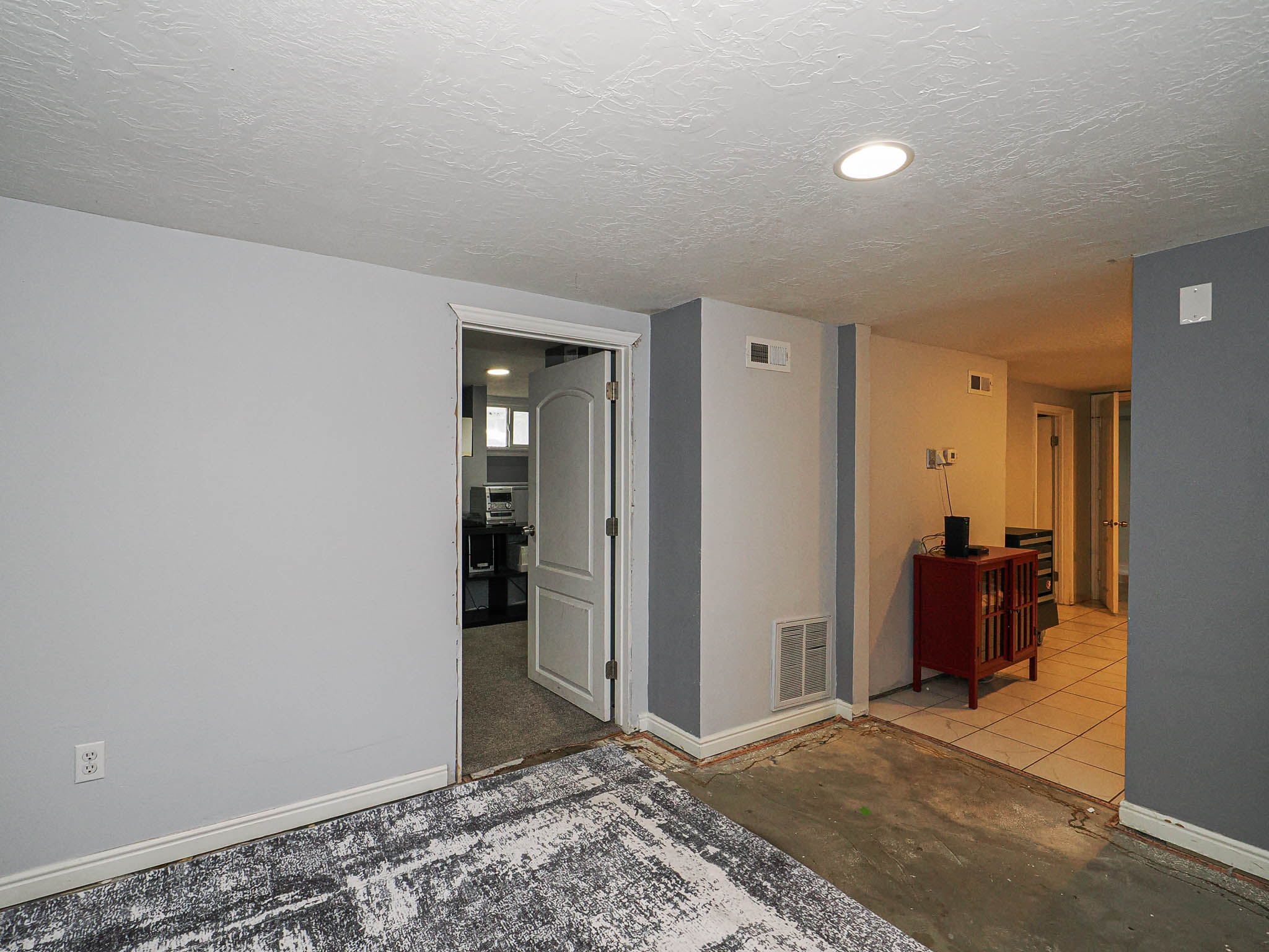 Recessed lighting illuminates a space featuring light gray walls, white baseboards, and a partial area with a patterned floor covering