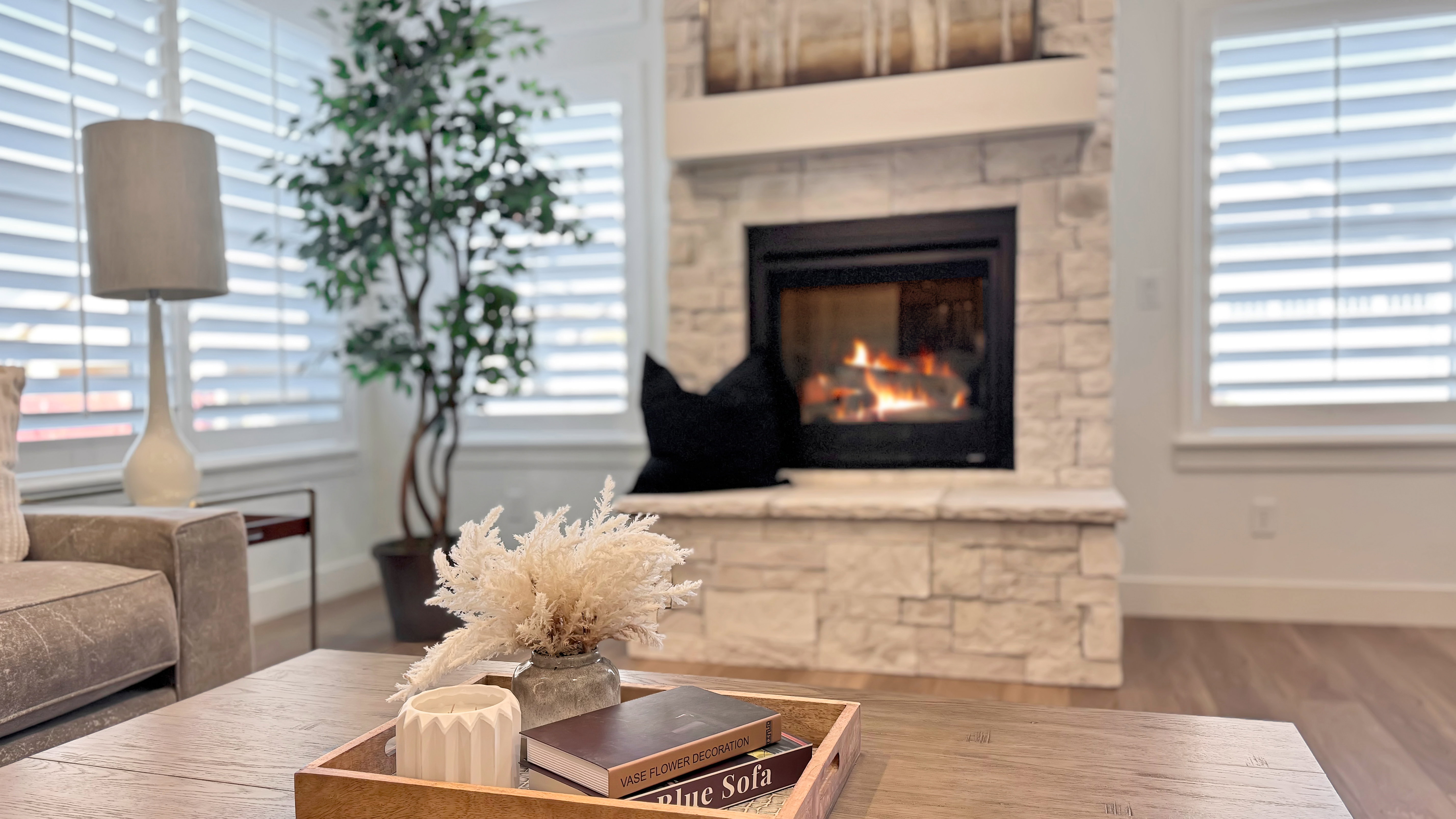 Stacked stone fireplace with a white mantel, wood-finish flooring, and white plantation shutters on multiple windows