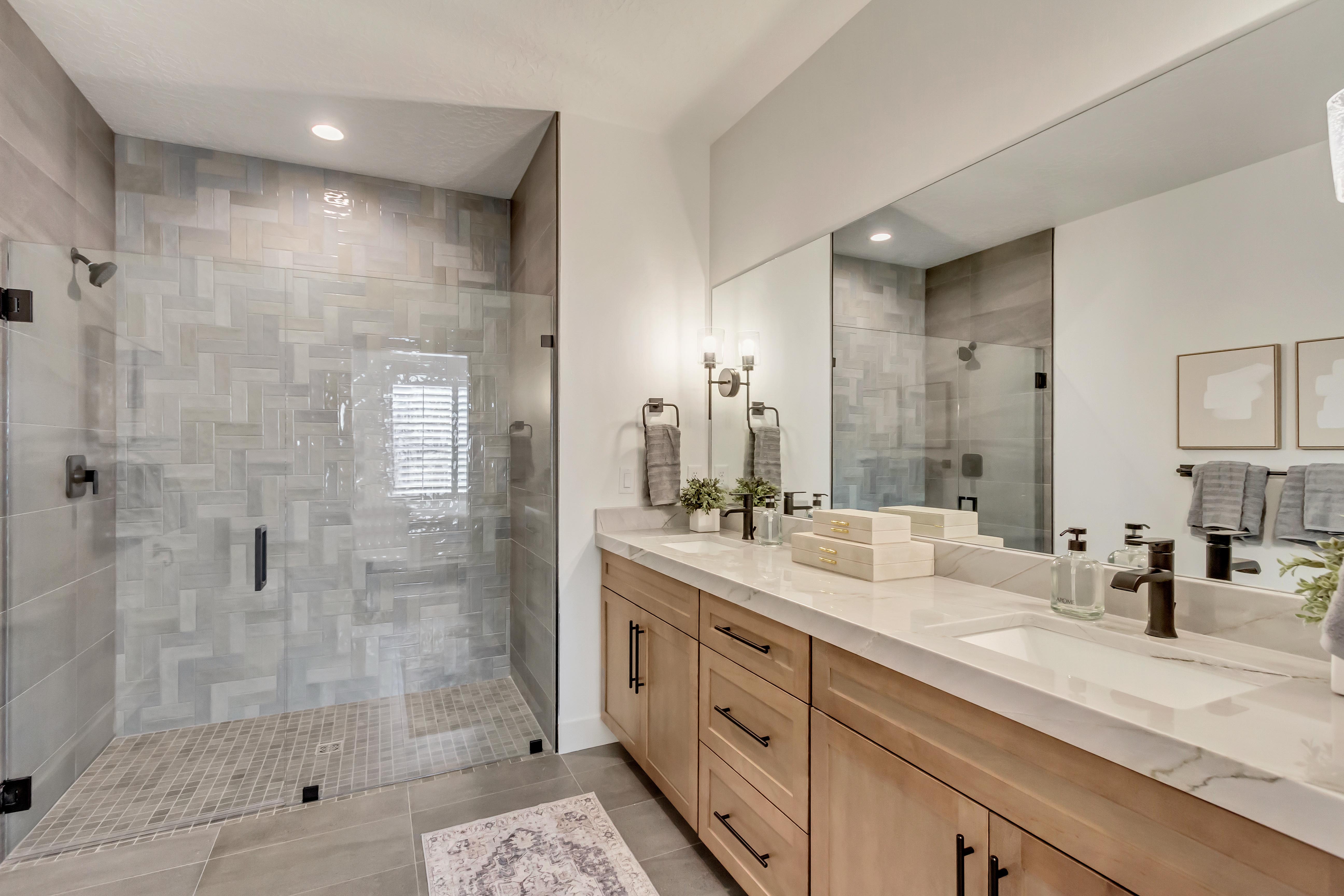 Spacious bathroom featuring a glass-enclosed shower with herringbone tile, a double vanity with light wood cabinetry, white countertops, and contemporary black fixtures