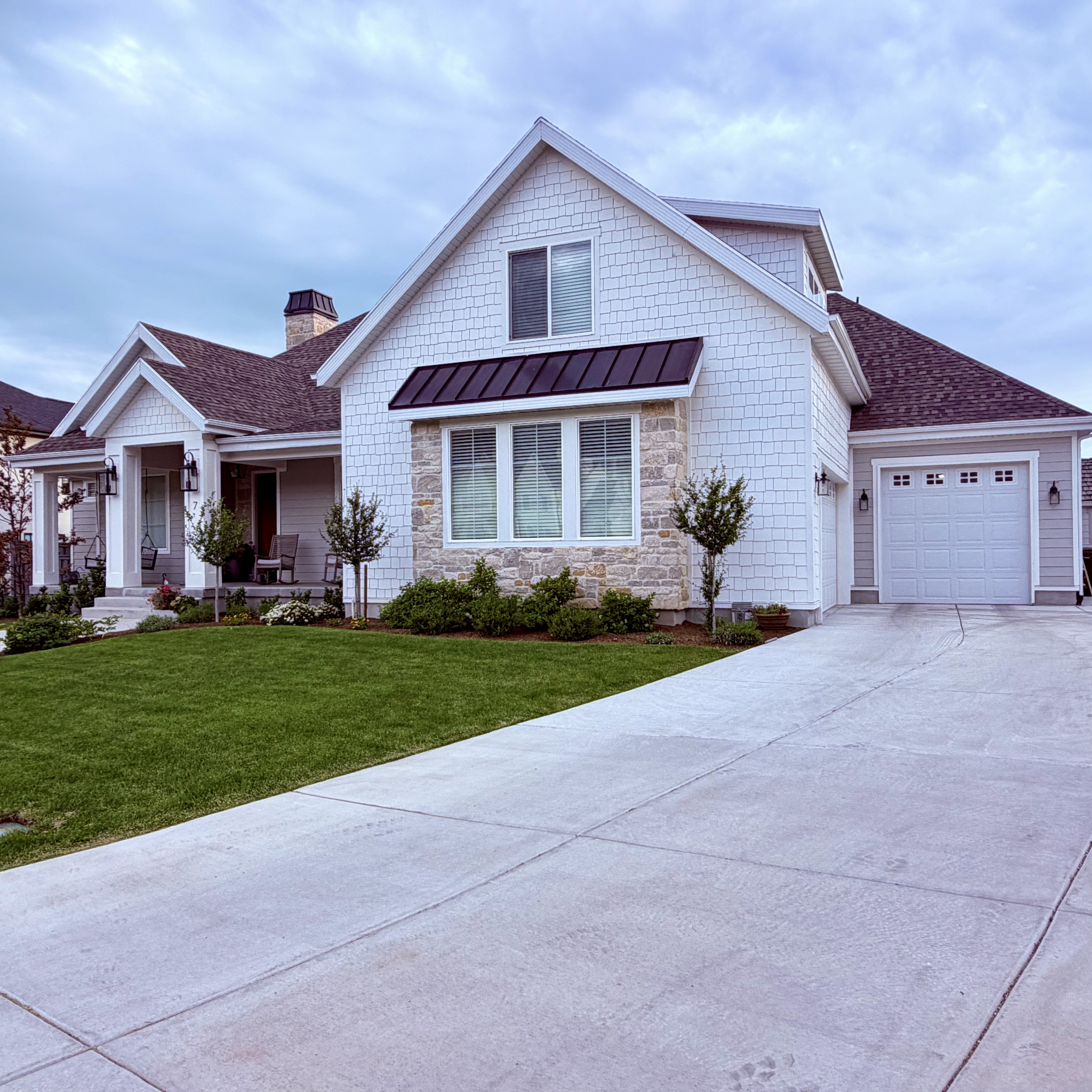 Inviting exterior featuring white shake siding, natural stone accents, a covered porch, an attached garage, and a concrete driveway