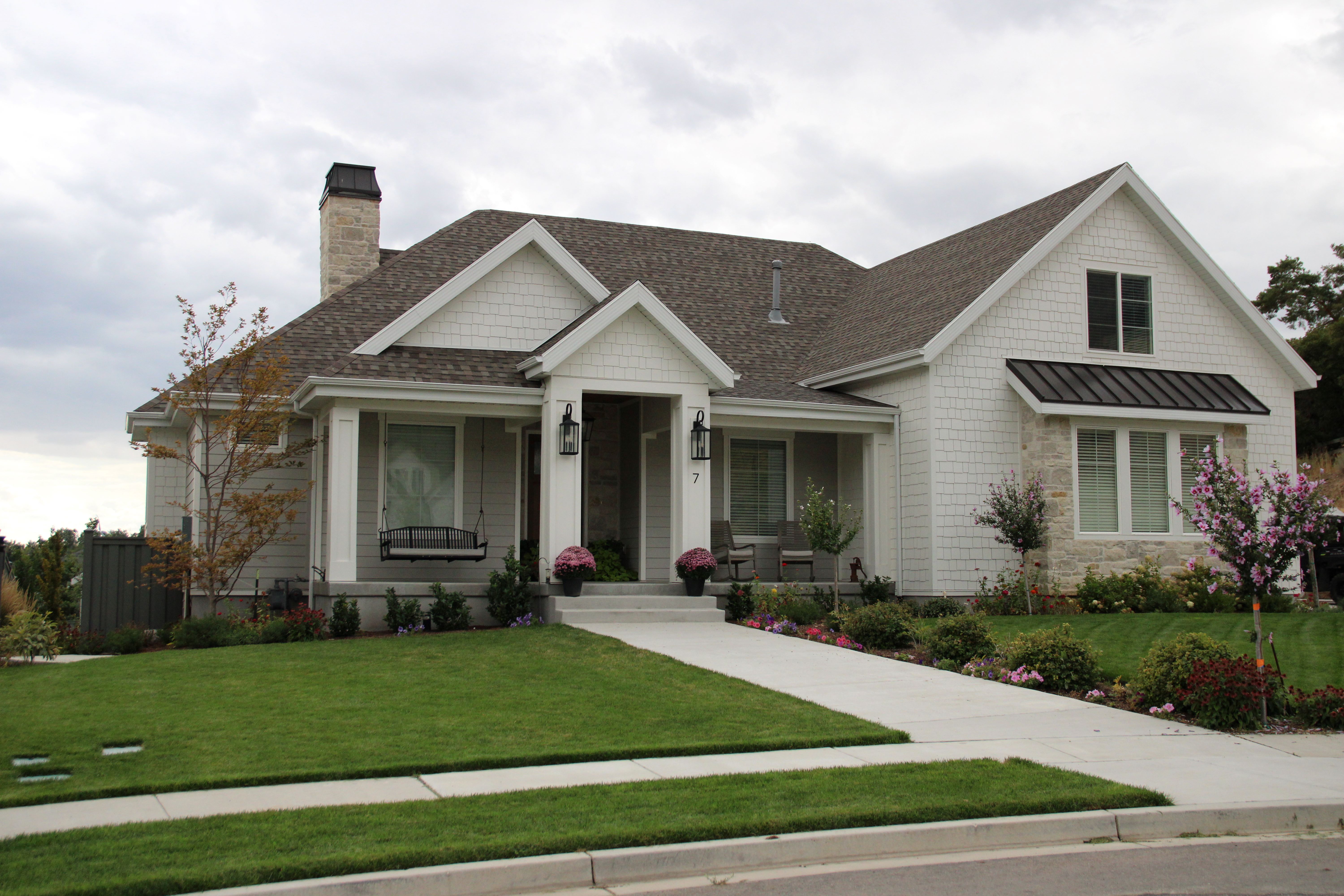 Craftsman-style home featuring a prominent front porch, mixed material facade with white shingle siding and stone accents, a dark shingle roof, and a meticulously landscaped front yard with a concrete walkway