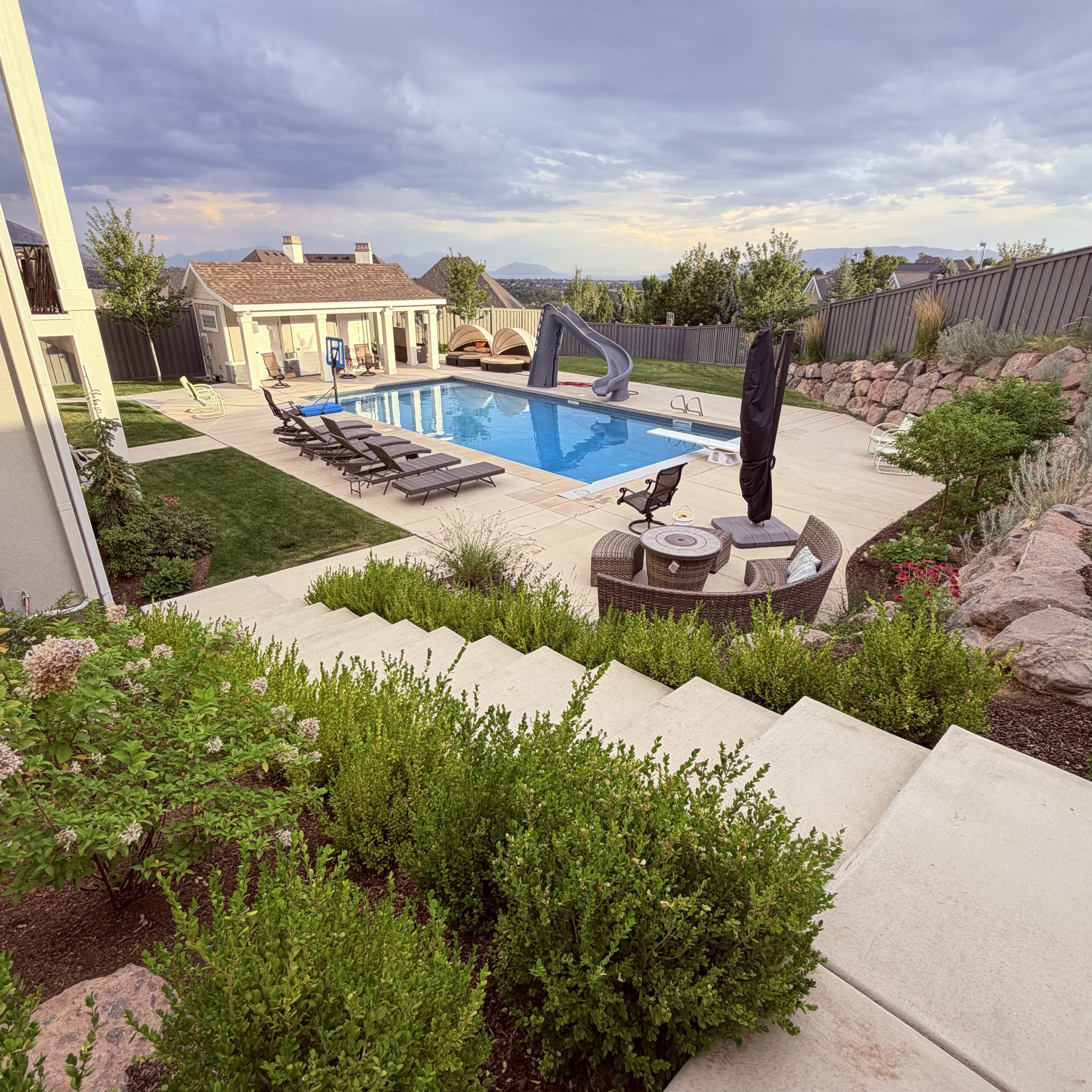 Spacious backyard featuring a rectangular swimming pool with integrated slide, a dedicated pool house, extensive concrete decking, and tiered landscaping with natural rock elements