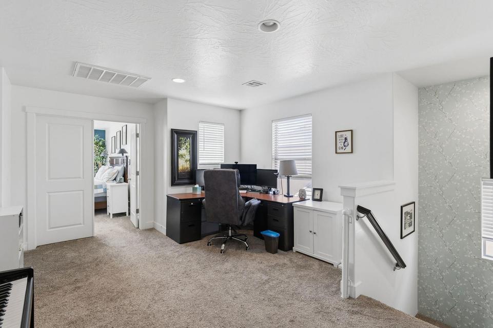 Carpeted upper-level space featuring a partial view of a bedroom through double pocket doors, a dedicated office area with a window, recessed lighting, and a staircase with a white balustrade and black handrail