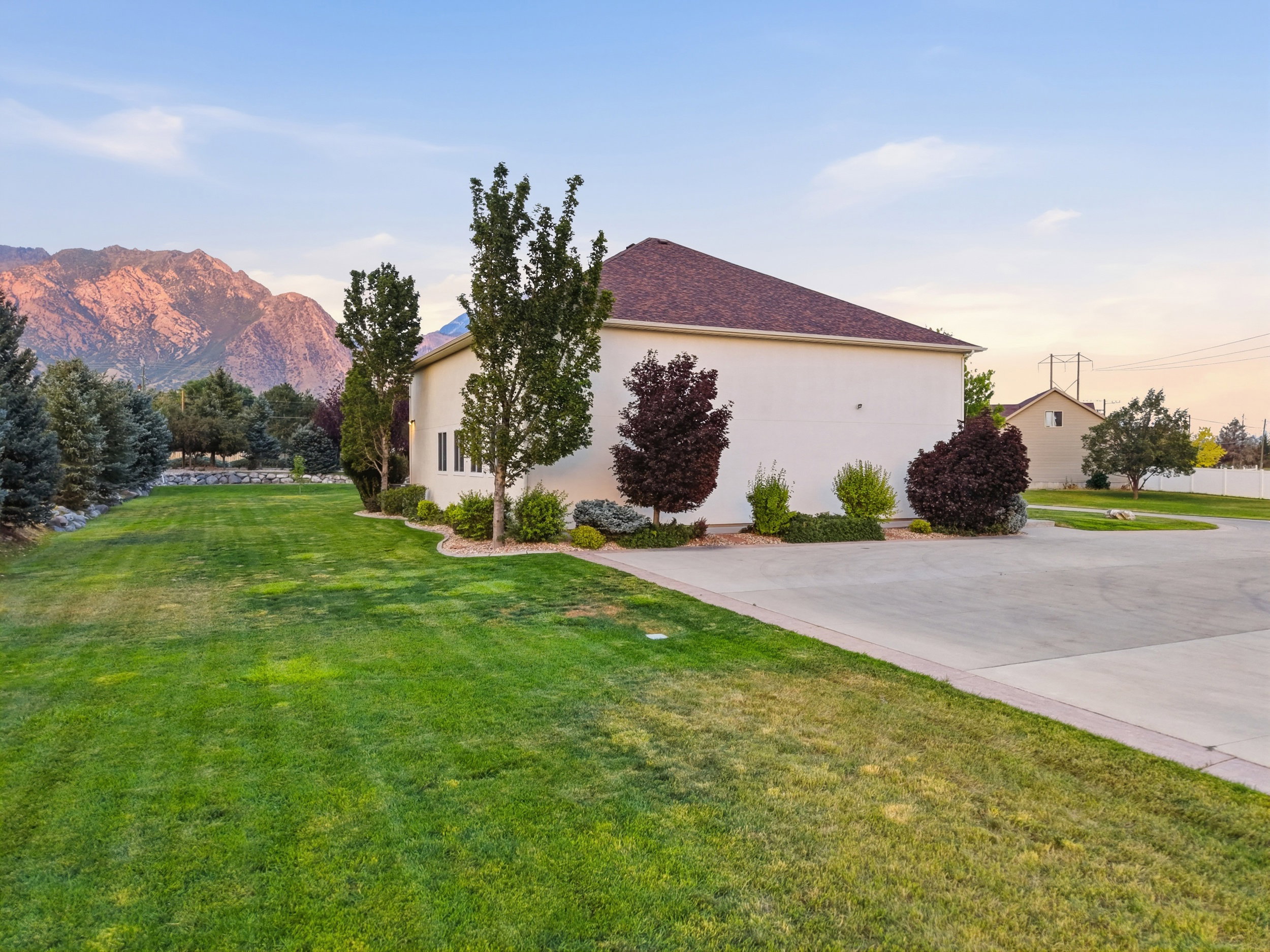 Spacious exterior featuring a light-colored facade, brown shingle roof, extensive green lawn, mature trees, and a concrete driveway