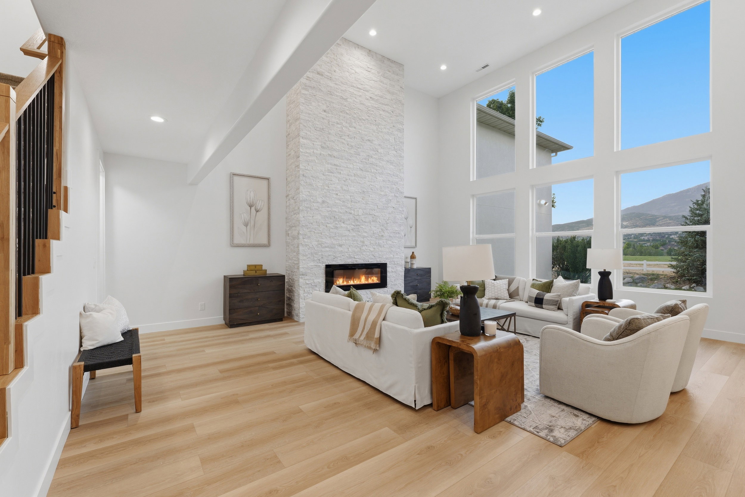 Spacious living area featuring expansive floor-to-ceiling windows, a tall stone-clad fireplace with a modern insert, light wood-finish flooring, recessed lighting, and a staircase with open wood treads and black metal balusters