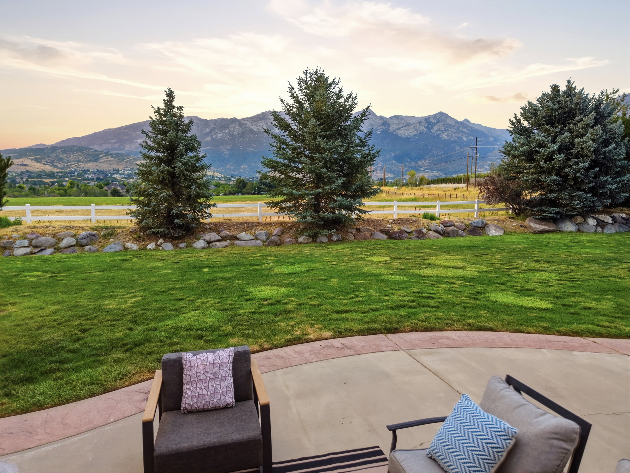 Expansive backyard featuring a large concrete patio with a decorative border, a manicured lawn, and mature evergreen trees