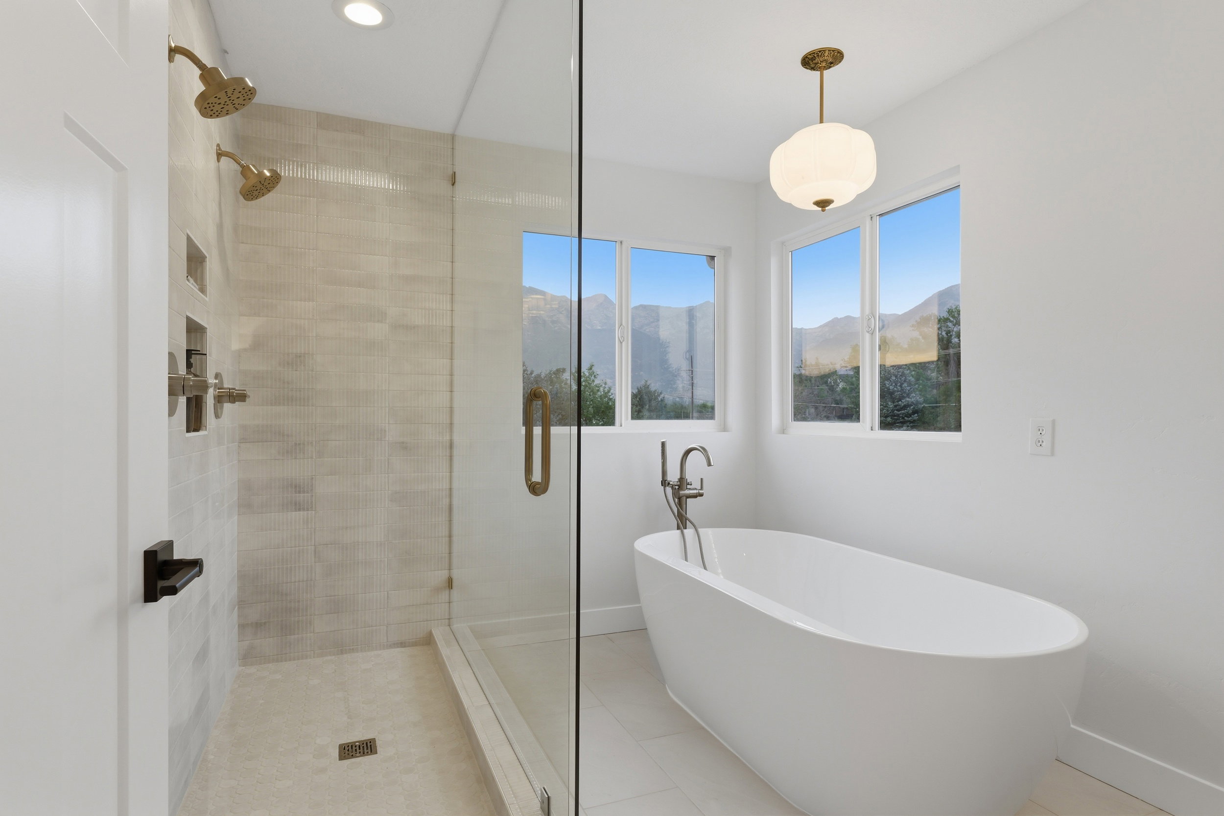 Spacious bathroom featuring a frameless glass shower enclosure, dual shower heads with gold-tone fixtures, a freestanding soaking tub, and multiple windows with mountain scenery