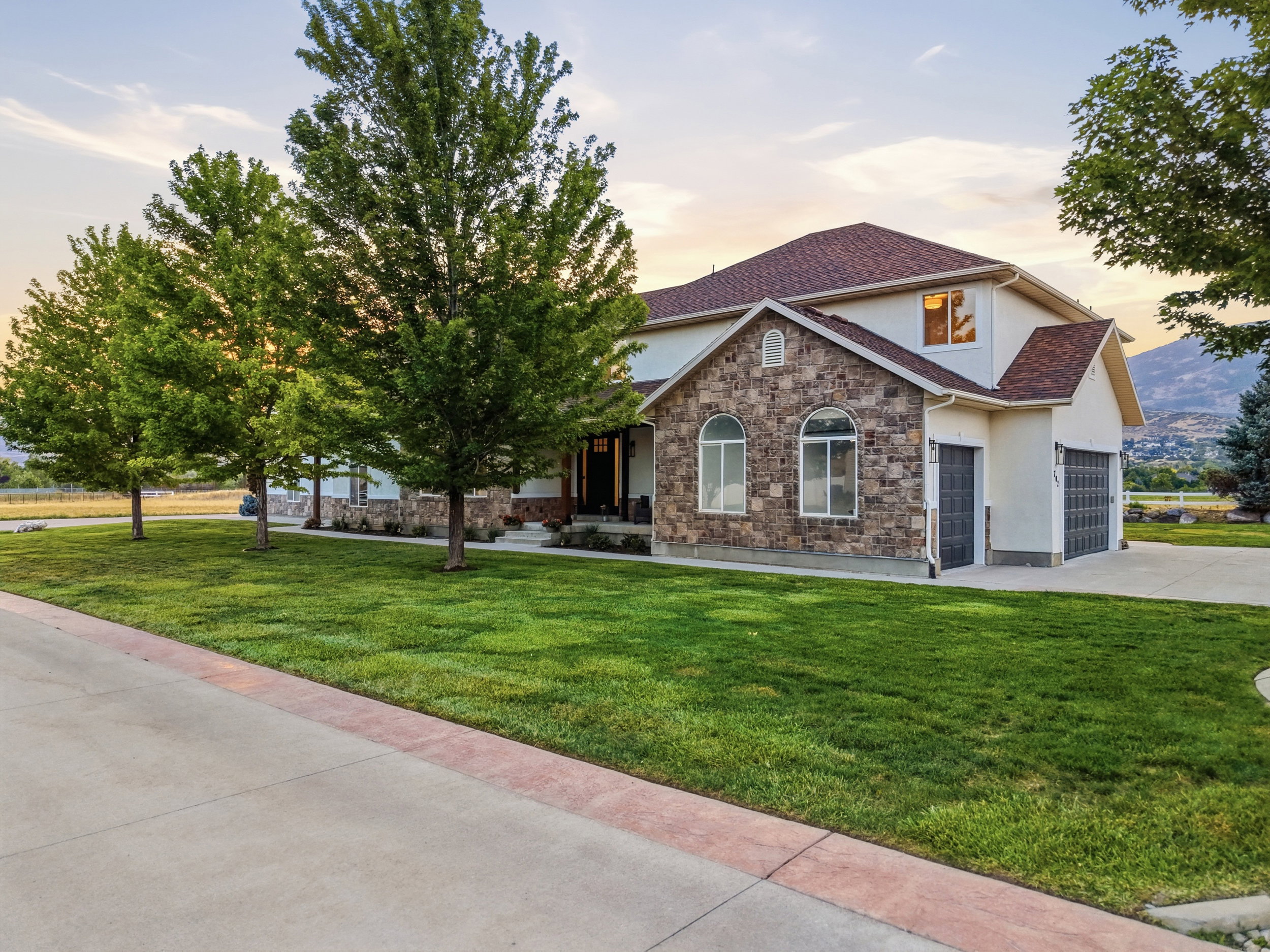 Stone and stucco exterior with an attached garage, featuring a concrete driveway and mature trees bordering a manicured lawn