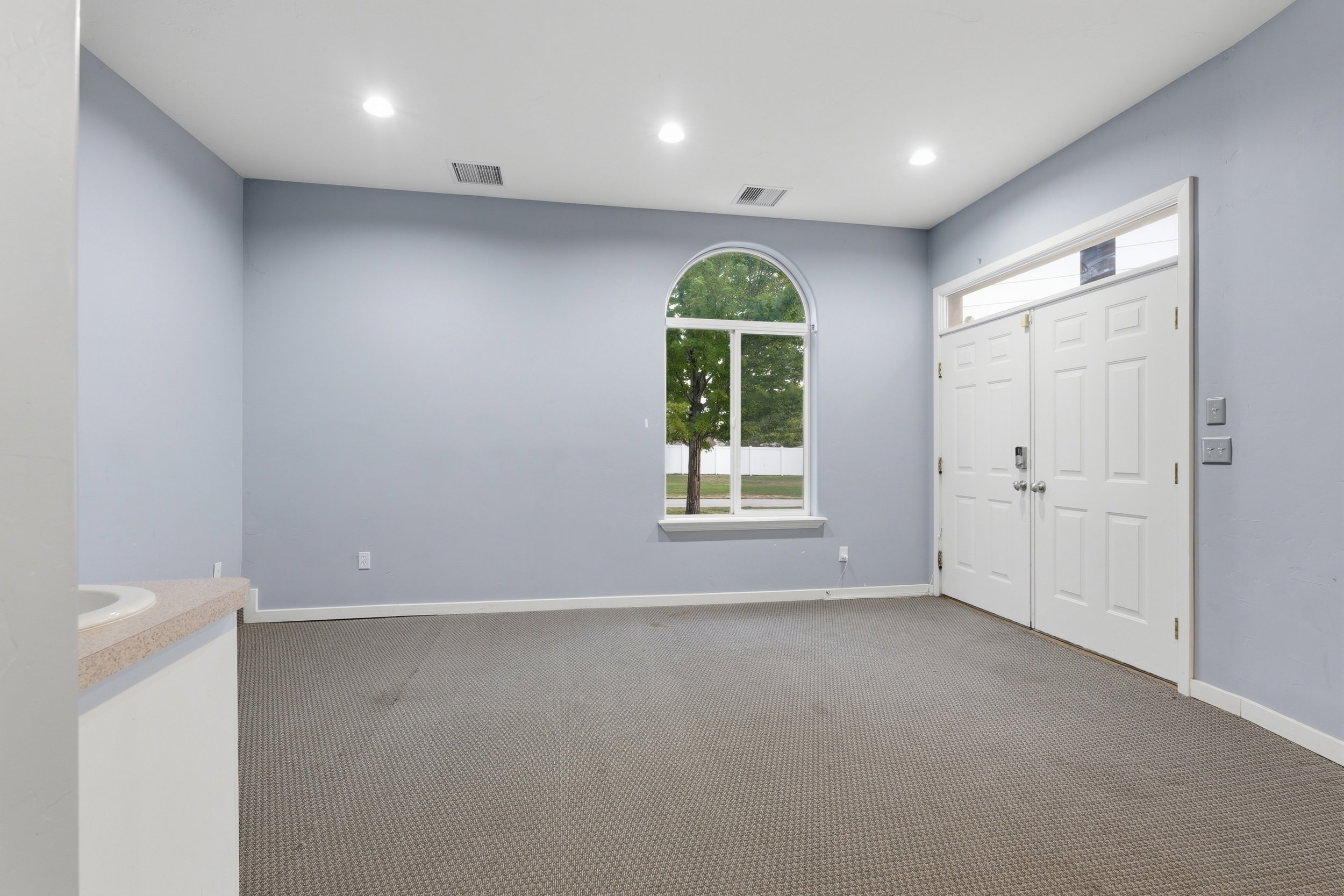 Spacious room featuring a prominent arched window, double entry doors with a transom window, recessed ceiling lighting, and light-colored carpeting