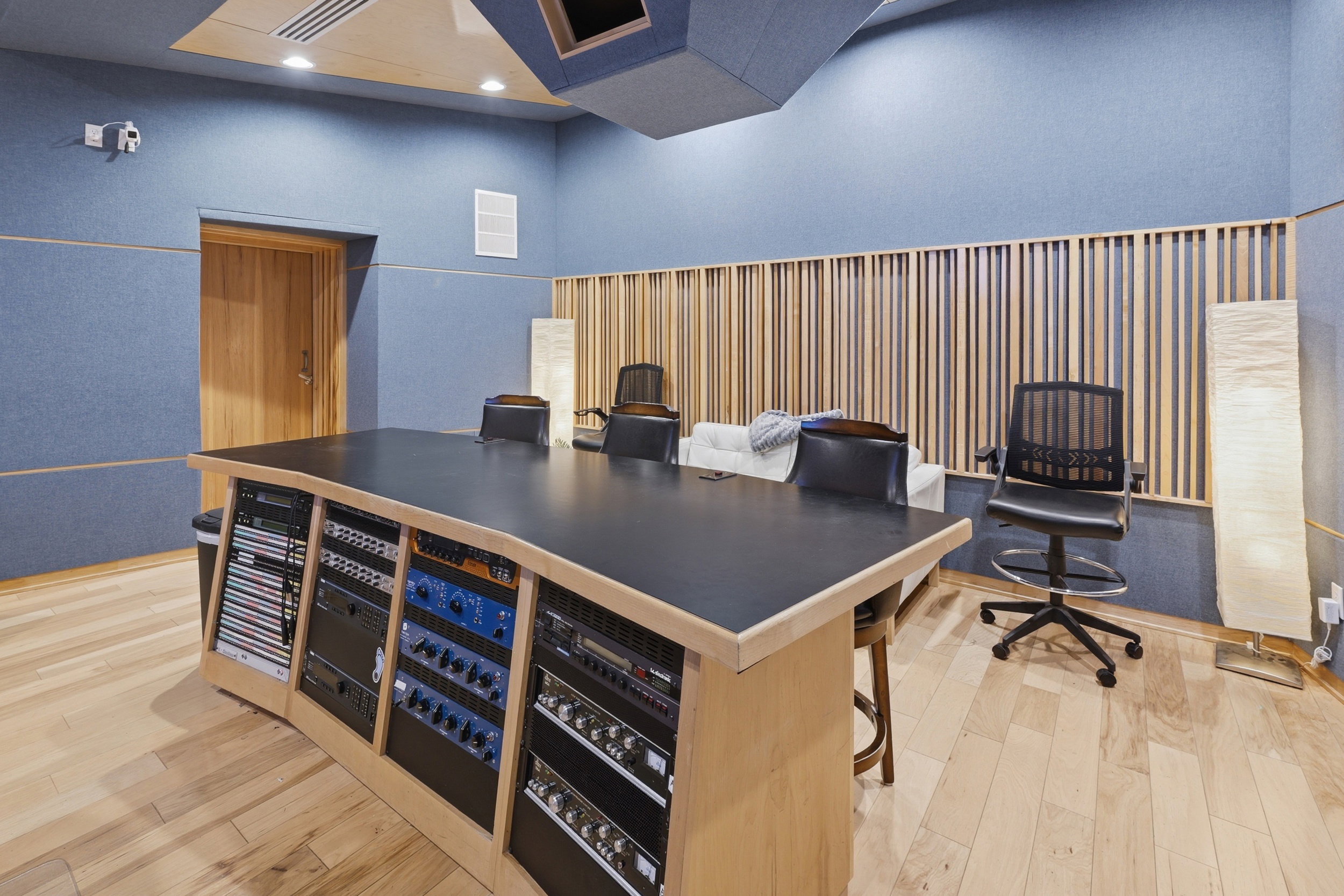 Professional studio space featuring a large built-in console with integrated equipment racks, acoustically treated walls, a natural wood-finish door, wood slat wall accents, and light wood-finish flooring