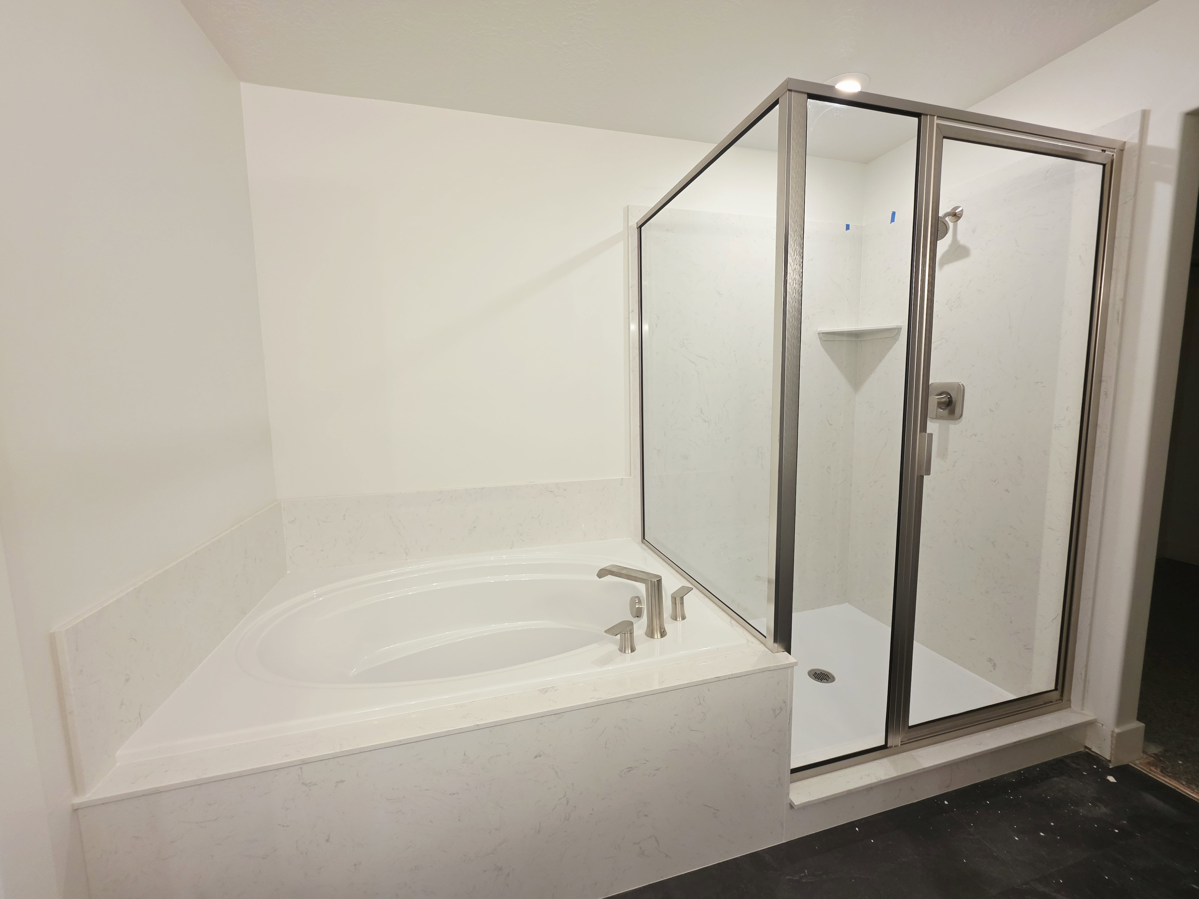 Integrated corner soaking tub with a brushed nickel faucet, set within a light-toned stone surround, complementing the adjacent glass-enclosed shower featuring a built-in shelf and brushed nickel fixtures