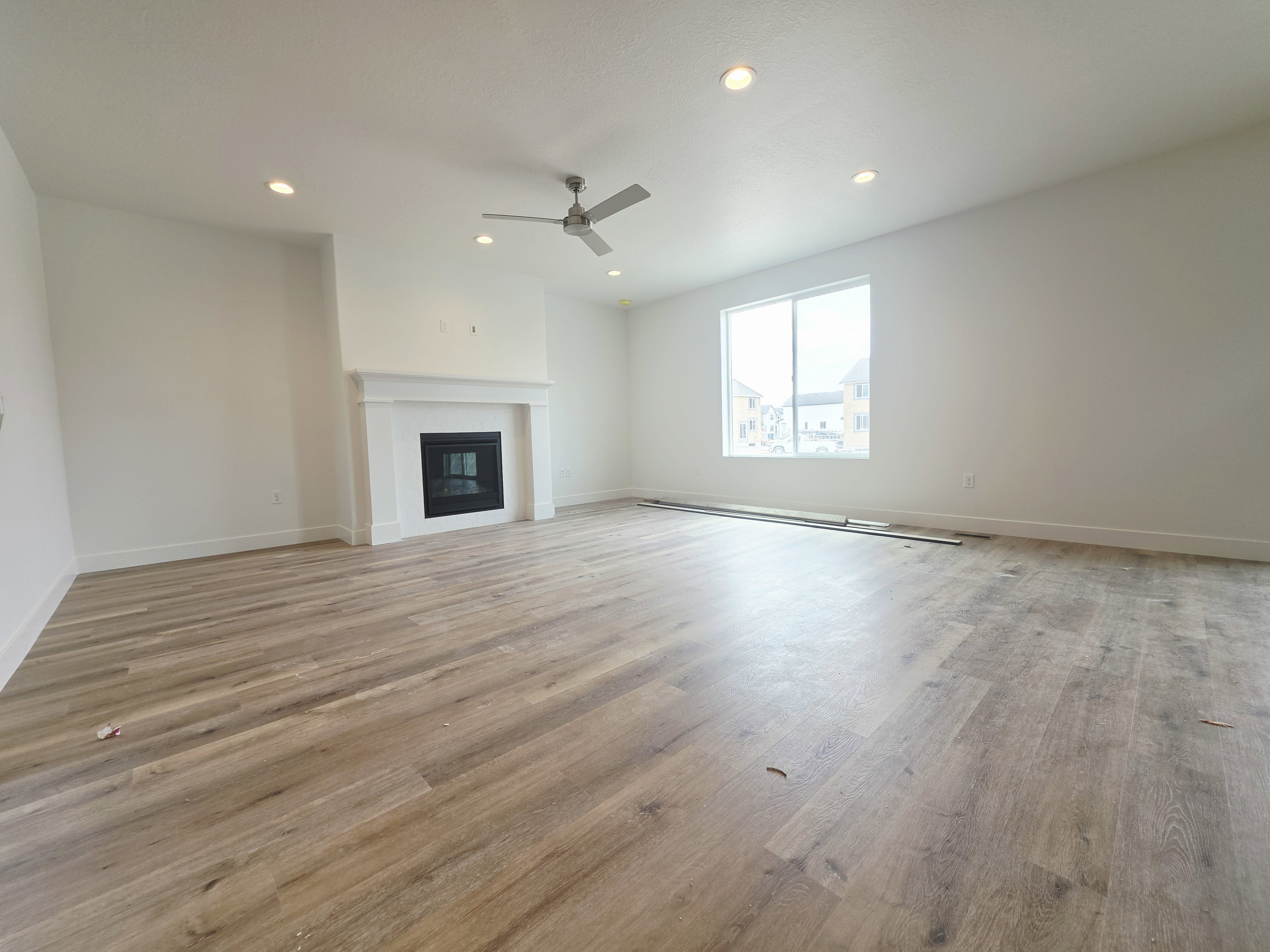 Spacious room featuring light wood-finish flooring, a white mantel fireplace, large window, ceiling fan, and recessed lighting