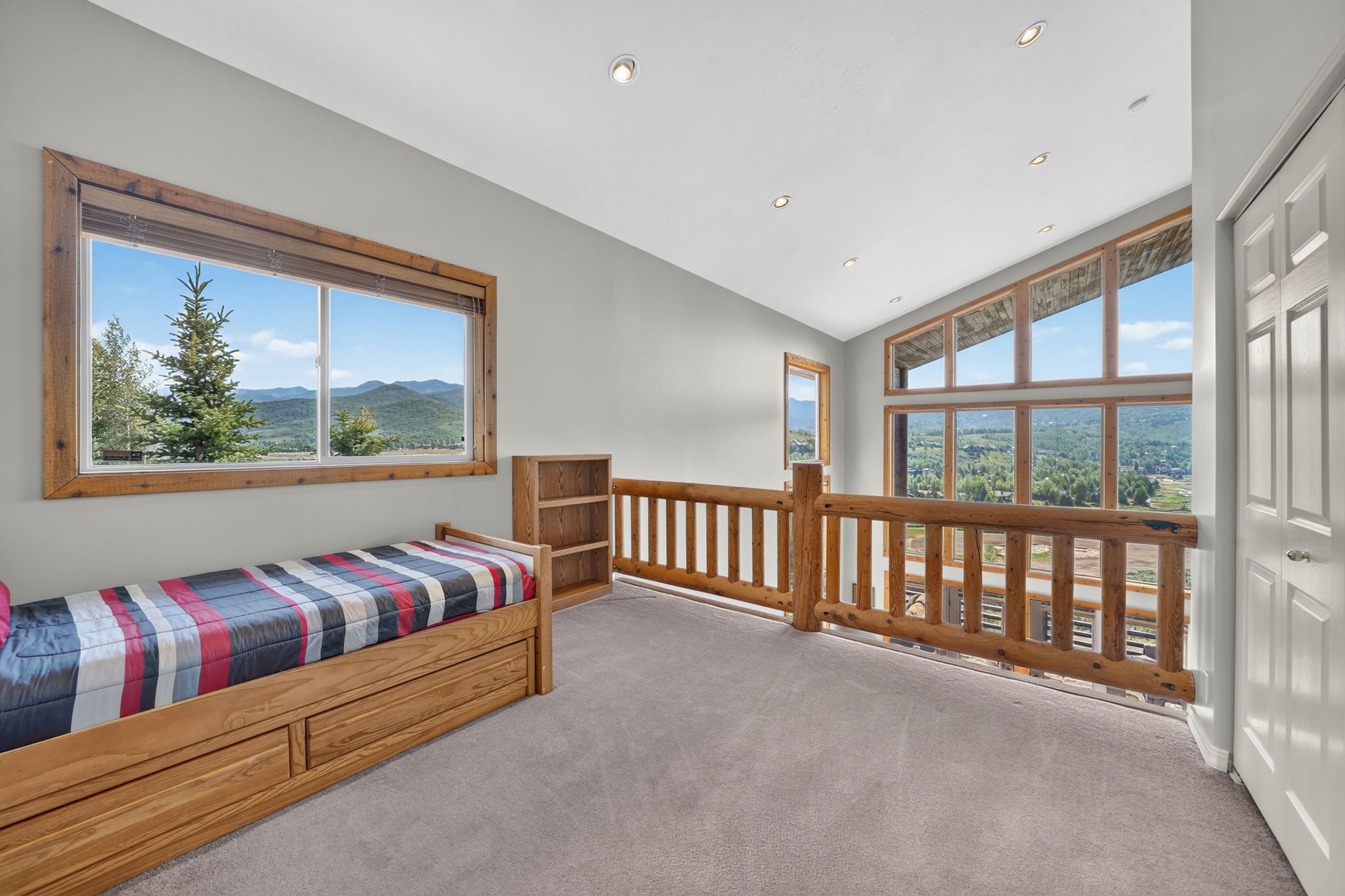 Upper-level loft space featuring a natural wood railing, light gray carpeting, and multiple large windows