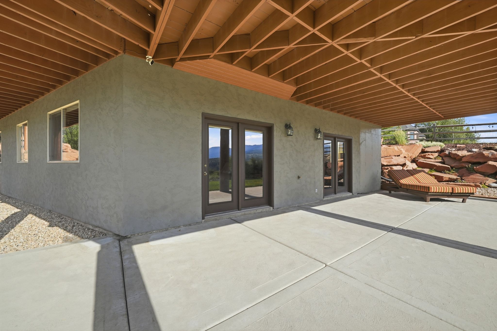 Expansive concrete patio with a full overhang, featuring multiple sets of glass-paned double doors and windows set into a textured exterior finish